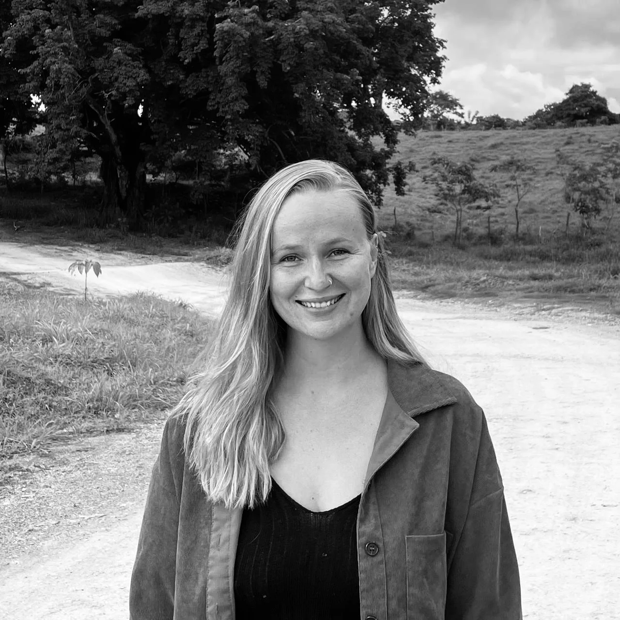 A smiling young woman with long hair, wearing a dark top and jacket, standing outdoors on a dirt path with trees and open land in the background.