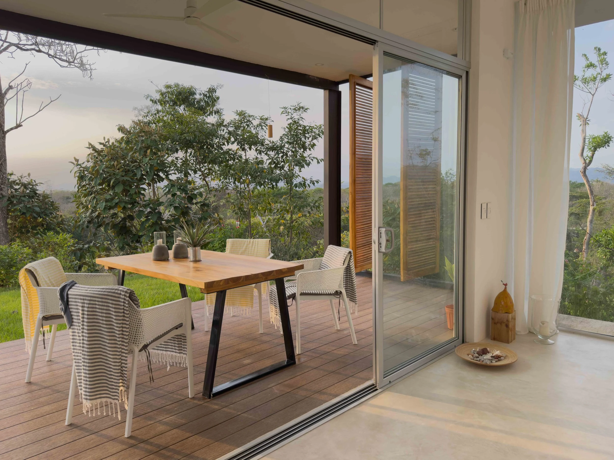 Indoor view of a glass sliding door leading to a wooden deck with a dining table and six chairs, overlooking greenery and trees, with natural daylight.