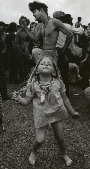 A young girl dancing outdoors with a confident expression, surrounded by a crowd of people, some of whom are shirtless and others wearing hats.