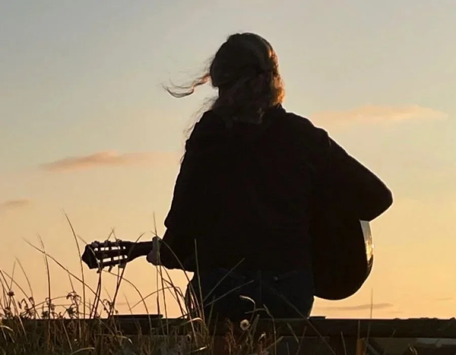 A person playing an acoustic guitar outdoors during sunset, viewed from behind, sitting among grass.