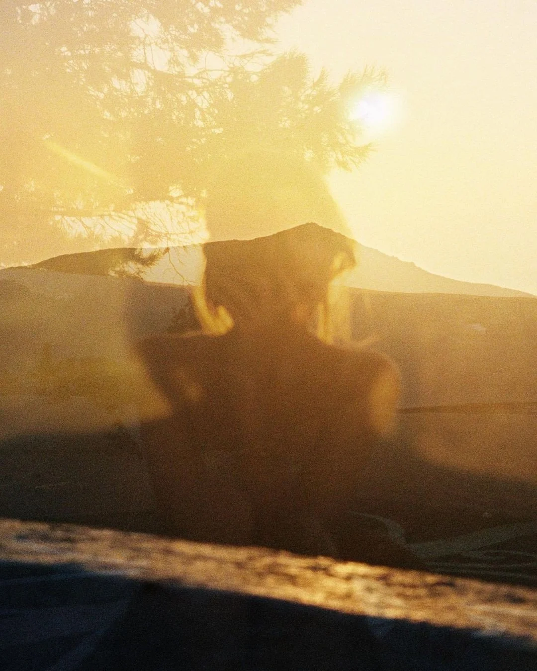 A reflection of a person in a car's windshield with a mountainous landscape and trees in the background, illuminated by sunlight.