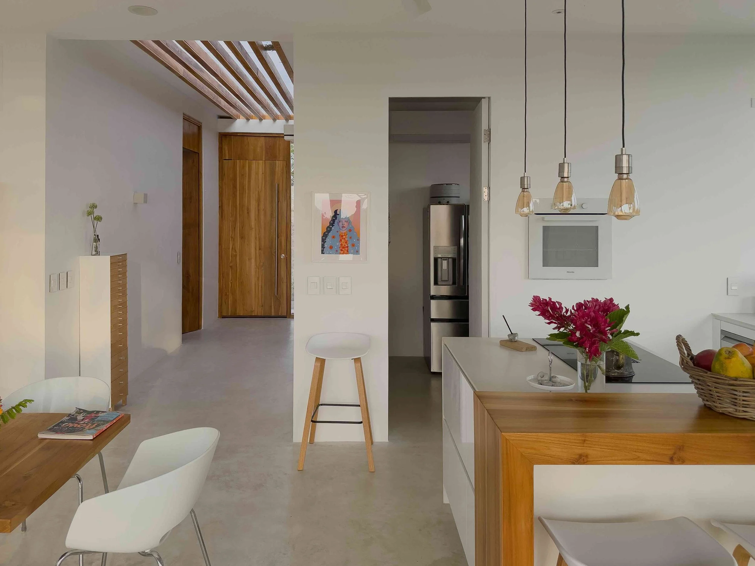 Modern kitchen and dining area with white walls, wooden accents, and minimal decor. There is a small table with white chairs, a kitchen island with a vase of pink flowers, and a basket of fruit. A doorway leads to a hallway with a wooden ceiling feat