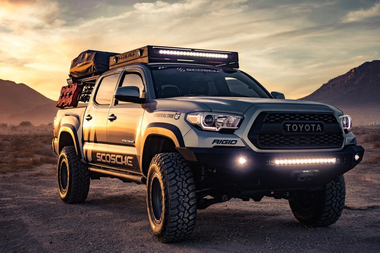 A modified Toyota Tacoma pickup truck with off-road tires, roof-mounted lights, and camping gear in a desert landscape during sunset.
