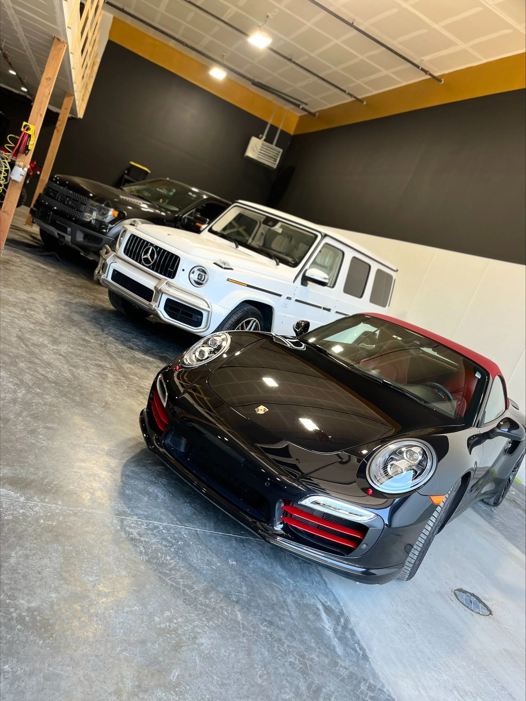 Showroom with four luxury cars: a black Porsche with red accents in the foreground, a white Mercedes-Benz G-Class SUV behind it, a black Chevrolet Silverado pickup truck, and another black vehicle partially visible on the far left. The floor is concr