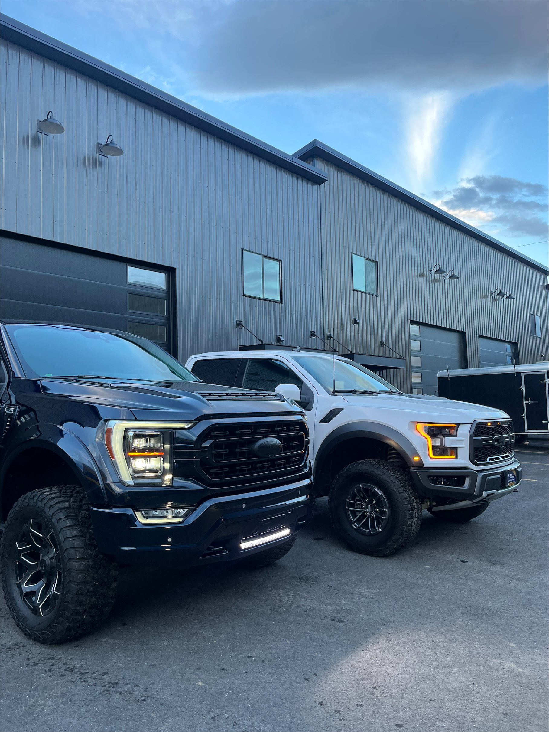 Two Ford trucks parked outside a large industrial building with metal siding, under a partly cloudy sky.