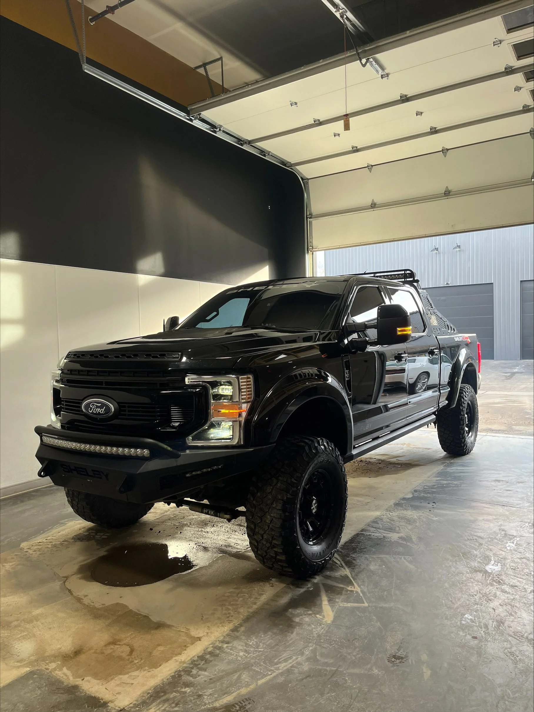 Black Ford pickup truck inside a garage with an open door, wet floor patches, and industrial surroundings.