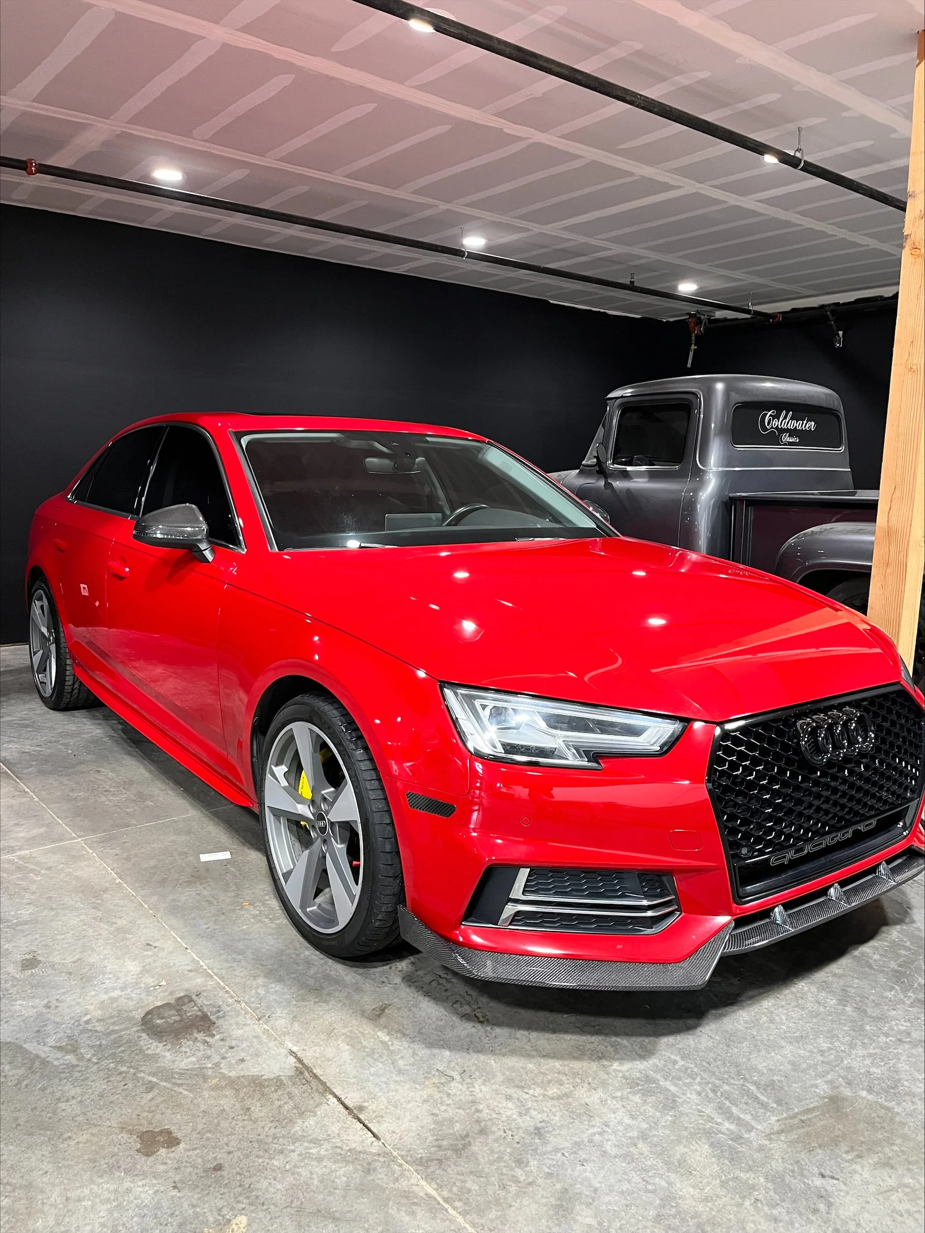 Red Audi sedan parked indoors with a black wall in the background and a vintage pickup truck behind it.