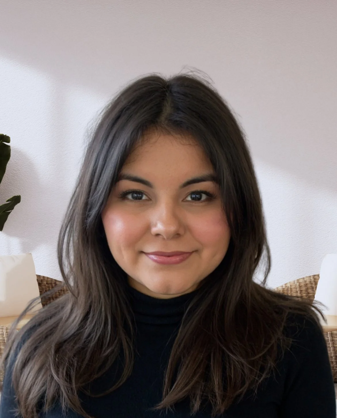 Portrait of a young woman with dark hair, wearing a black top, smiling at the camera. Background includes a light-colored wall and some green plant leaves.
