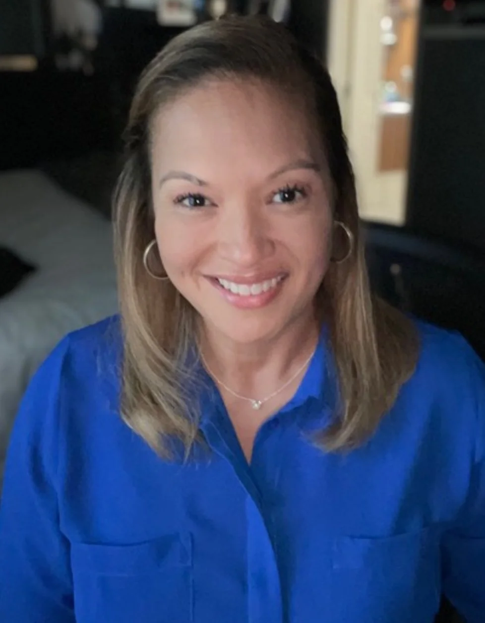 A woman with shoulder-length light brown hair, wearing a blue shirt, silver hoop earrings, and a delicate necklace, smiling at the camera in an indoor setting.