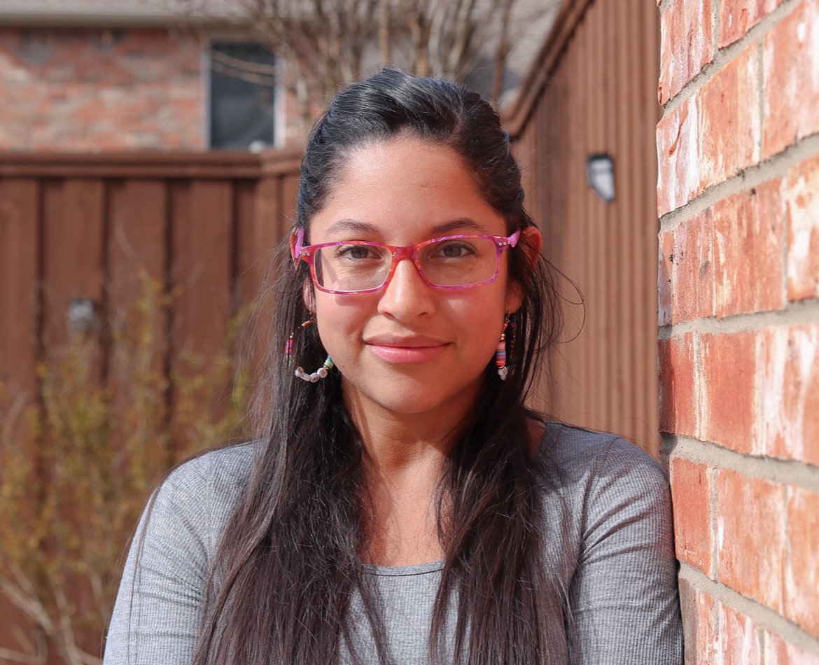 A woman with long dark hair, pink glasses, and colorful earrings leans against a brick wall in an outdoor setting.