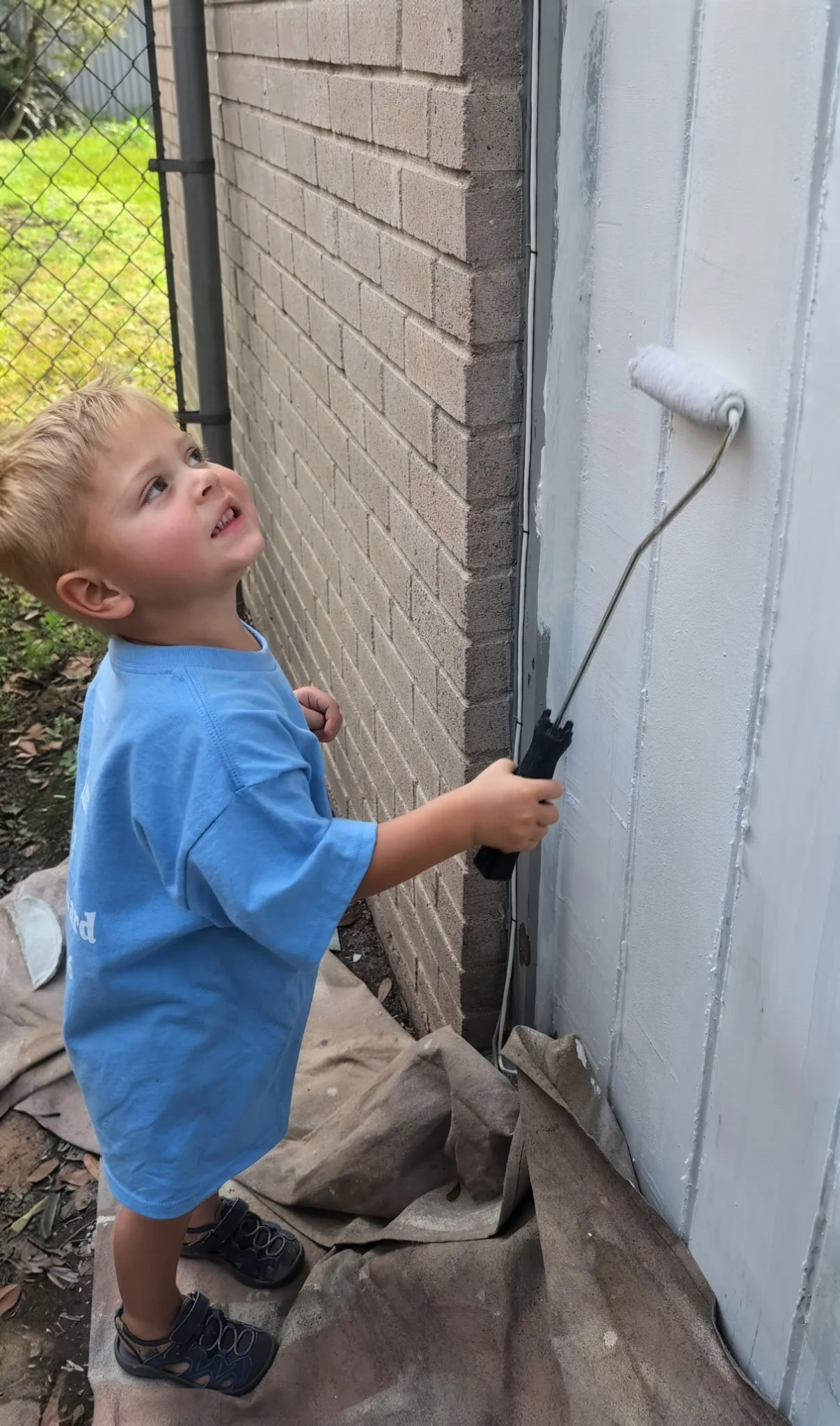 Toddler Isaac painting the exterior trim of Gayle’s home in Sugar Land during a Paint It Forward community project, supervised by Chris and his crew