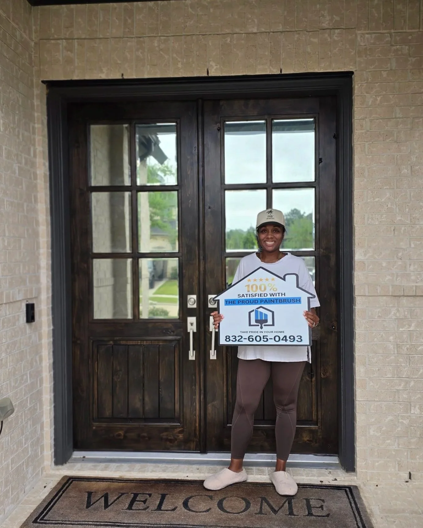 Happy homeowner standing in front of refinished dark walnut double front door in Katy, Texas holding 100 percent satisfied sign from The Proud Paintbrush