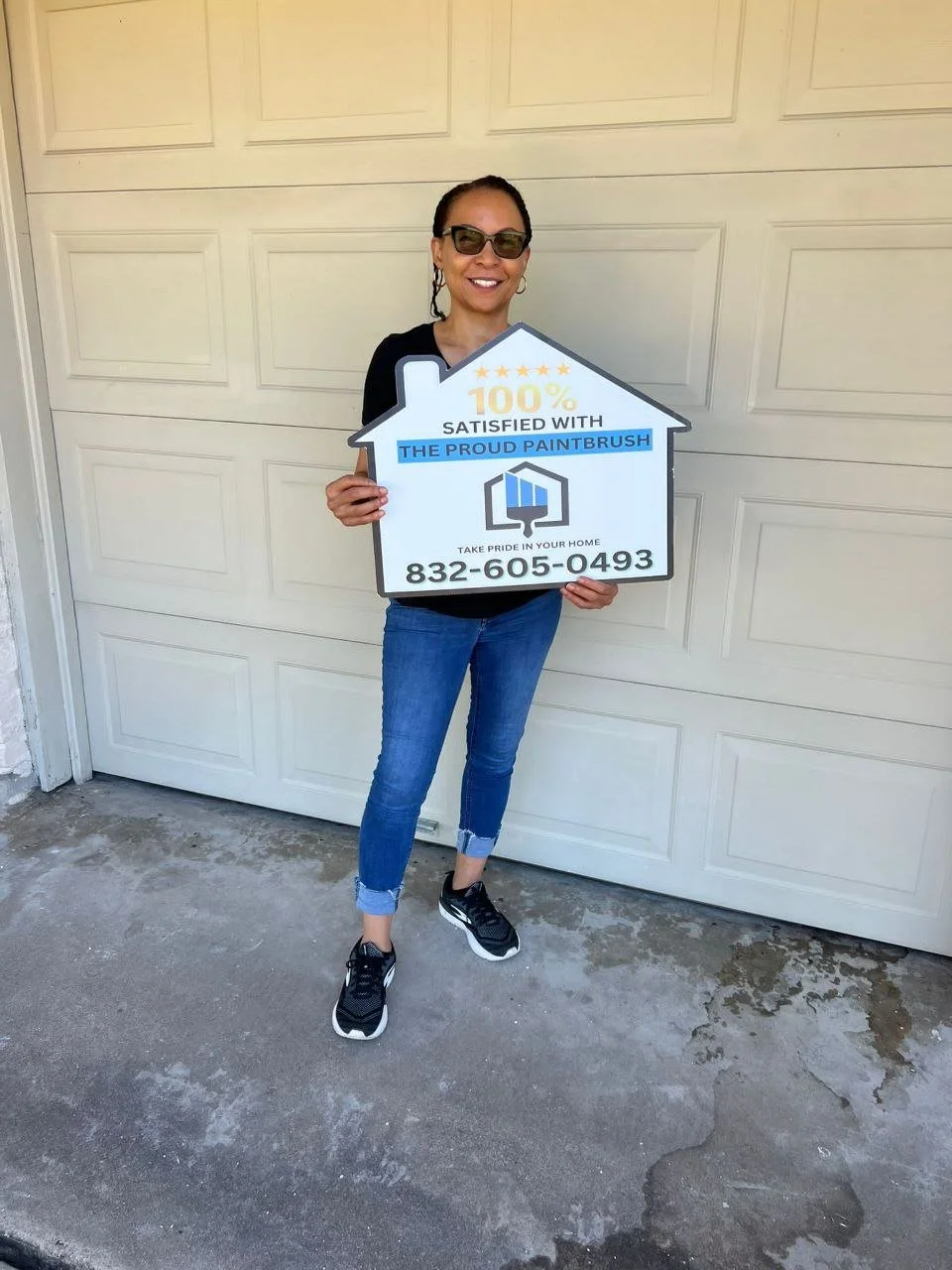 Woman wearing sunglasses holding a house-shaped sign that says 100% satisfied with The Proud Paintbrush, including a phone number.