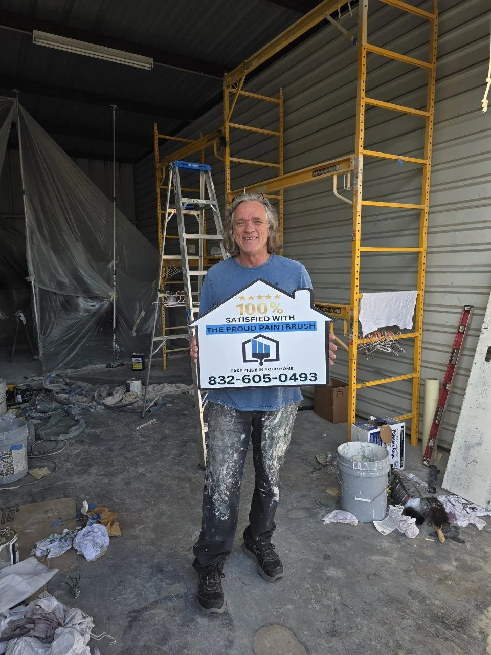 A man standing in a workshop or construction area, holding a house-shaped sign that reads '100% satisfied with the proud paintbrush,' with a phone number below. The background includes ladders, scaffolding, paint supplies, and construction debris.