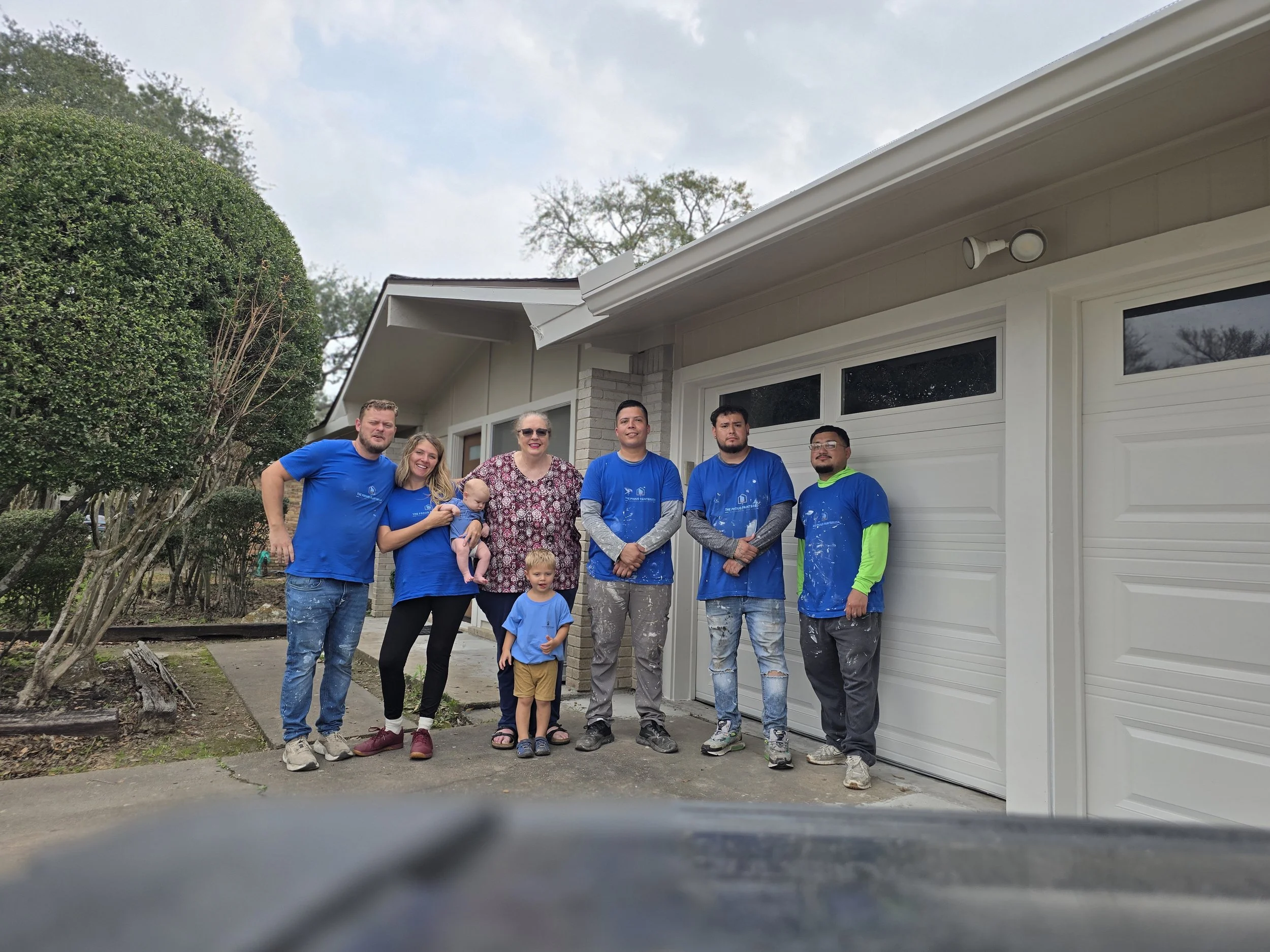 Three workers in blue shirts painting the exterior of Gayle’s house in Sugar Land with Chris, his wife Sarah, and children Sammy and Isaac watching and helping
