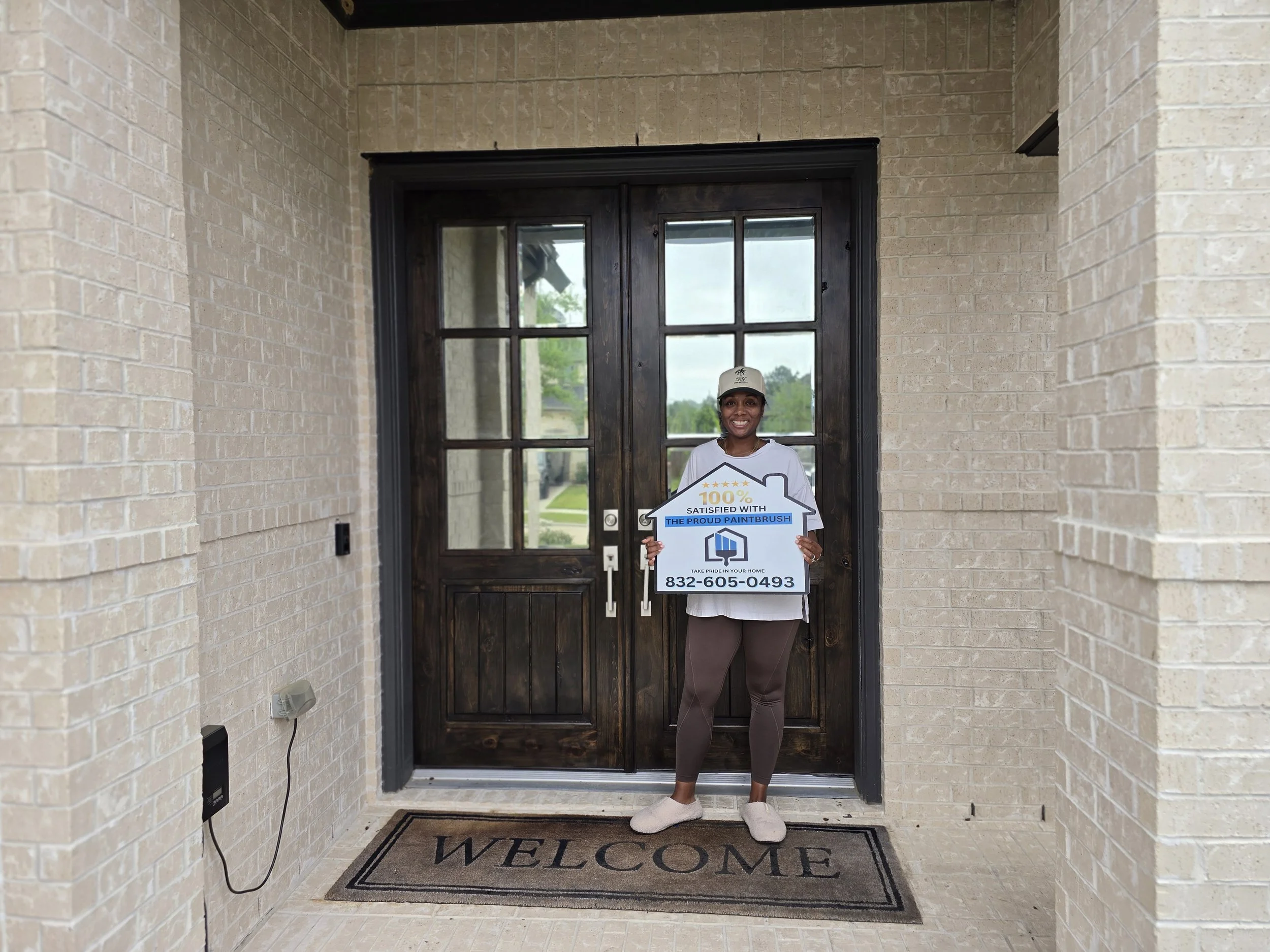 Happy homeowner standing in front of refinished dark walnut double front door in Katy, Texas holding 100 percent satisfied sign from The Proud Paintbrush