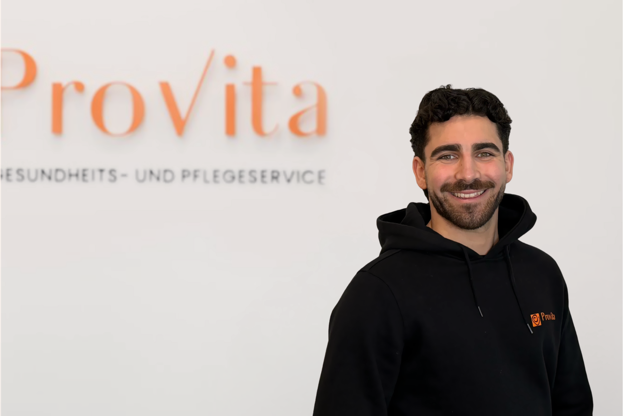 Ein Mann mit dunklen, lockigen Haaren und Schnurrbart, der vor einer weißen Wand mit dem Logo "Provita" und der Aufschrift "Gesundheits- und Pflegeservice" steht. Er trägt ein schwarzes T-Shirt mit einem kleinen Provita-Logo.