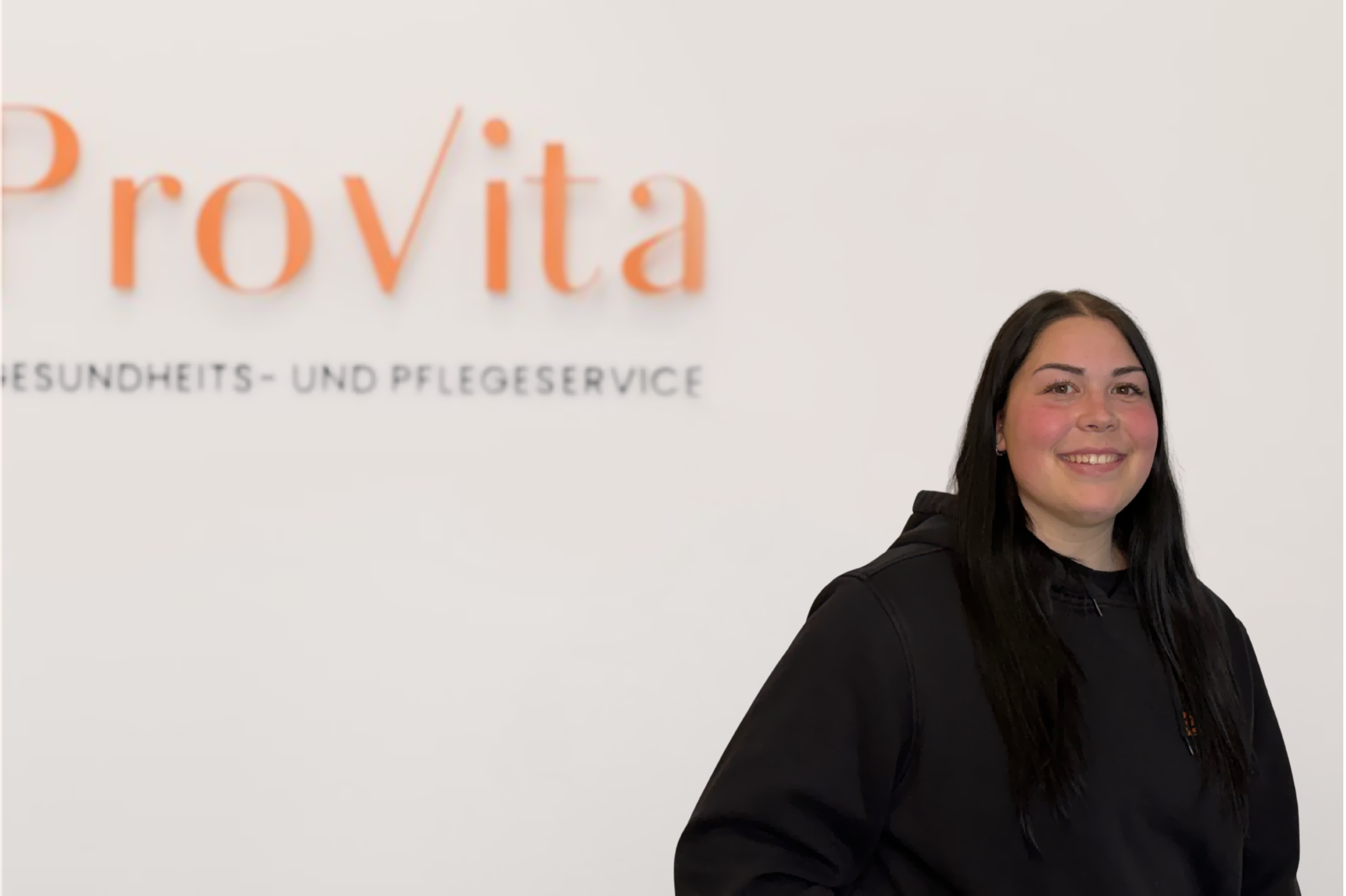 Mitarbeiterin des Pflegedienstes Provita lächelt selbstbewusst vor dem Logo- engagiert, herzlich und mit Freude im Einsatz für die Pflege.