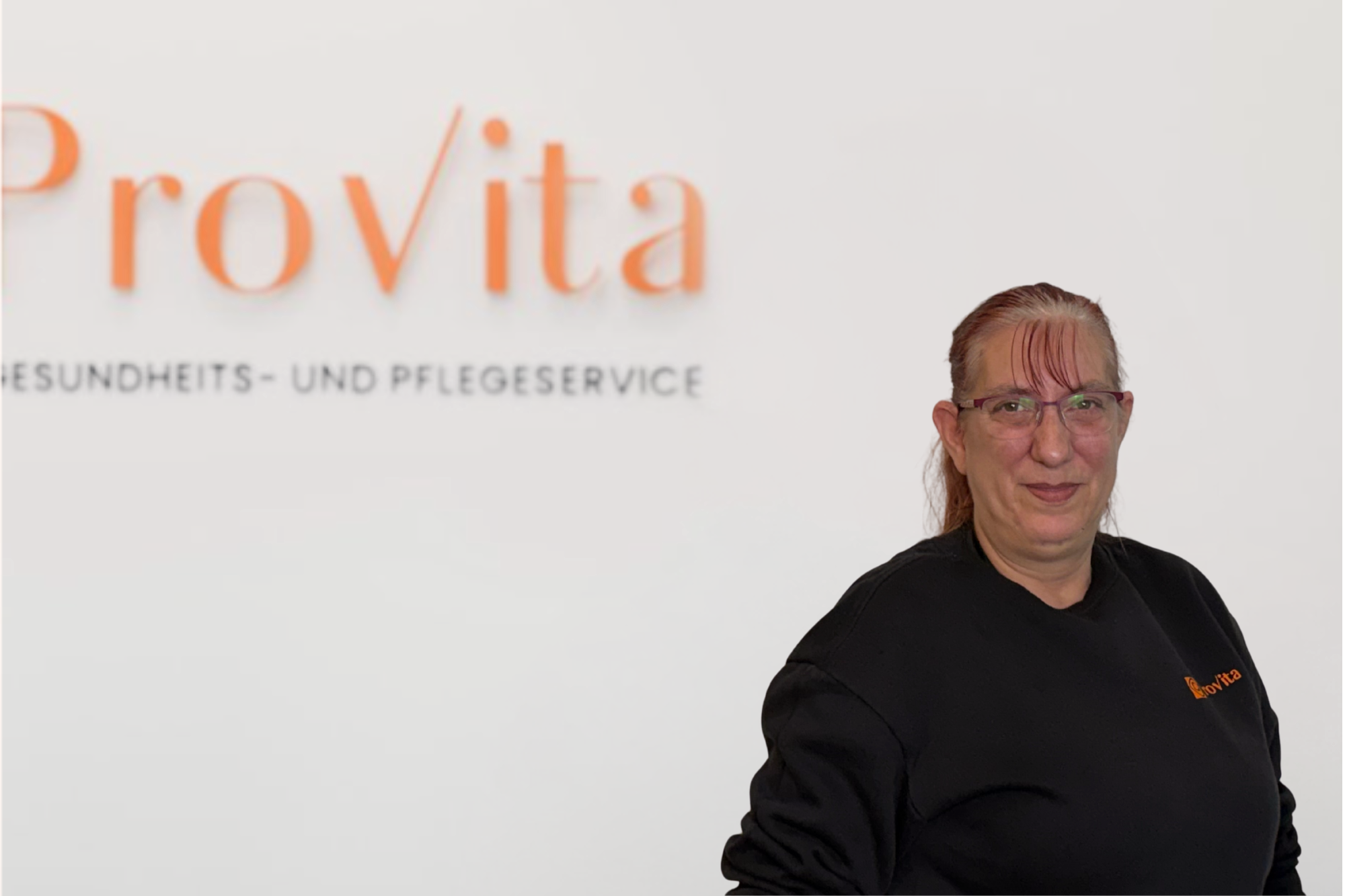 Frau mit Brille und schwarzen Hoodie vor weißer Wand mit dem Logo 'ProVita' und der Beschriftung 'Gesundheits- und Pflegeservice'.