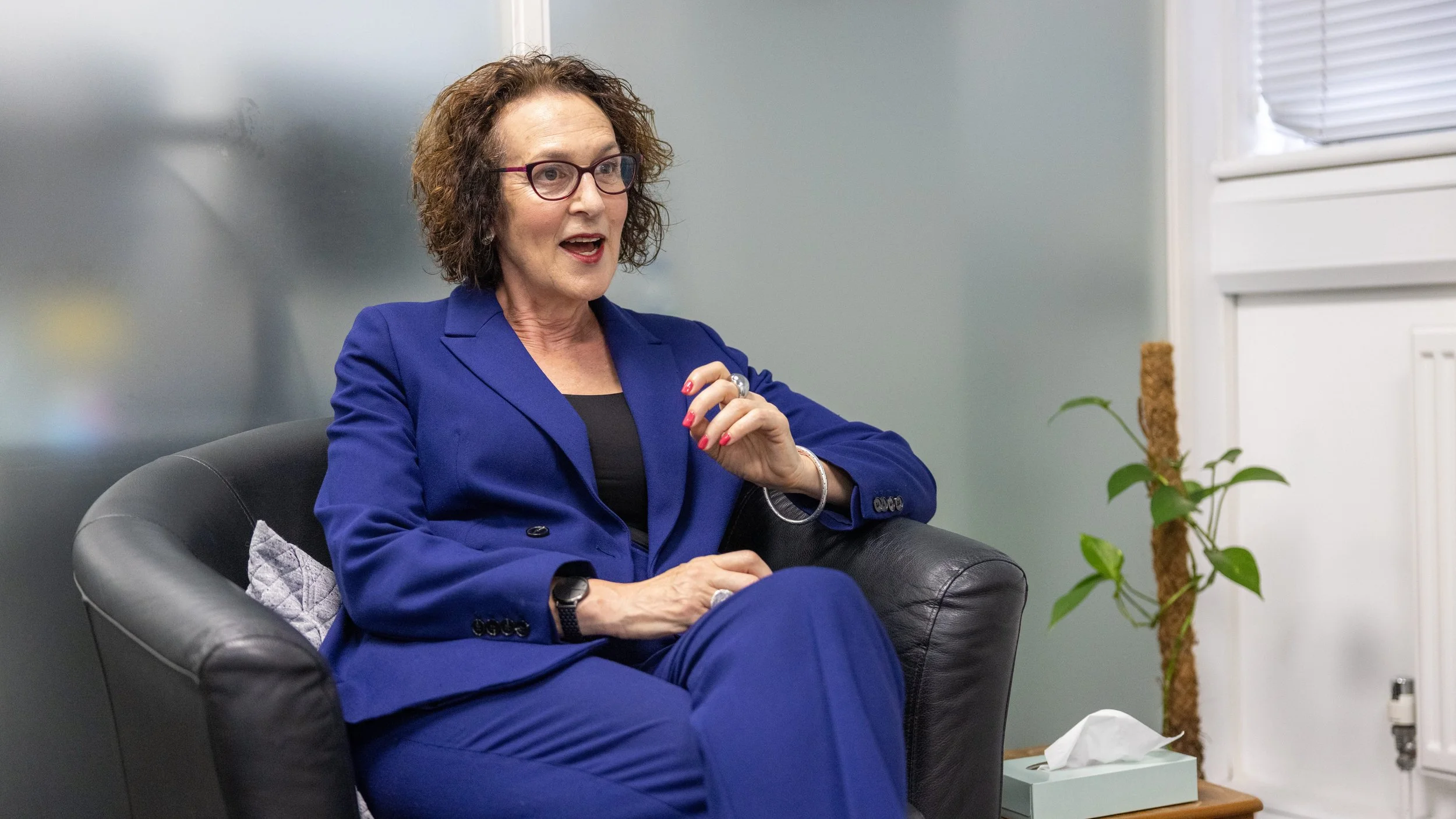 Three women sitting in a brightly lit room, engaged in conversation. They are seated on black armchairs in a semi-circle, with a green plant in the background. One woman wears a colorful scarf and glasses.