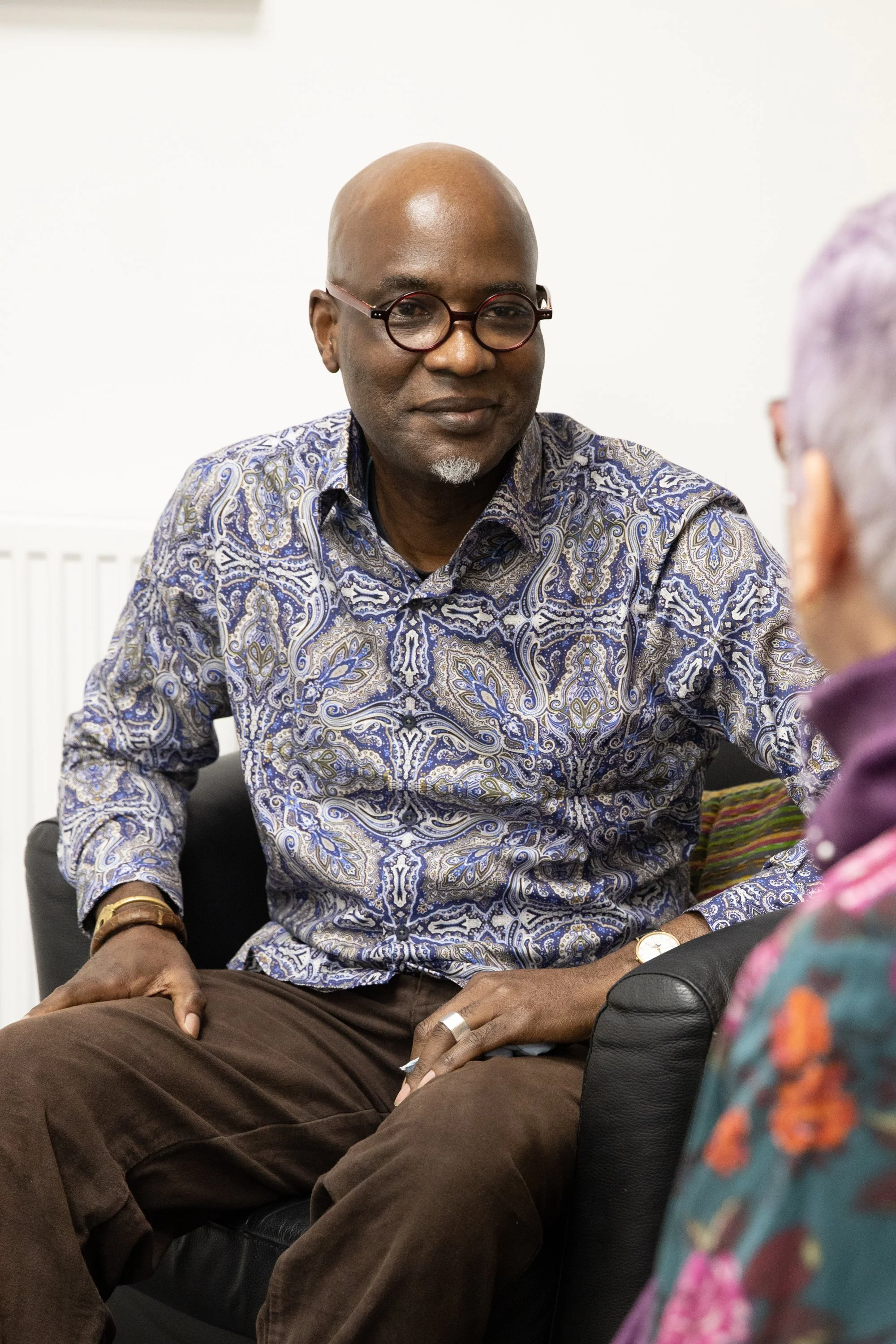 A man with glasses and a blue patterned shirt speaking with a woman with her back to the camera.
