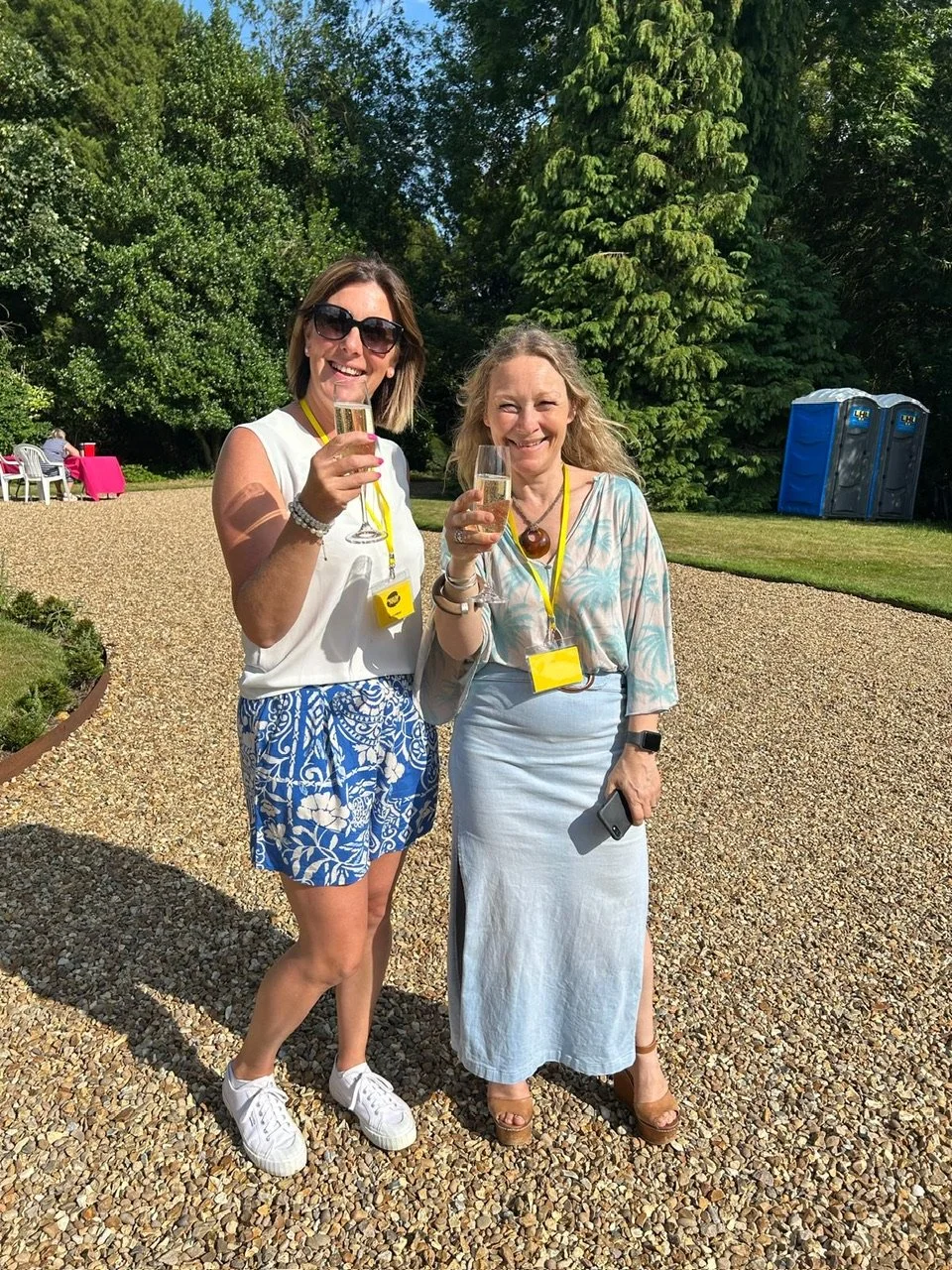 Two volunteers standing outdoors at Histon Manor, smiling, holding champagne glasses, dressed in summer clothes.