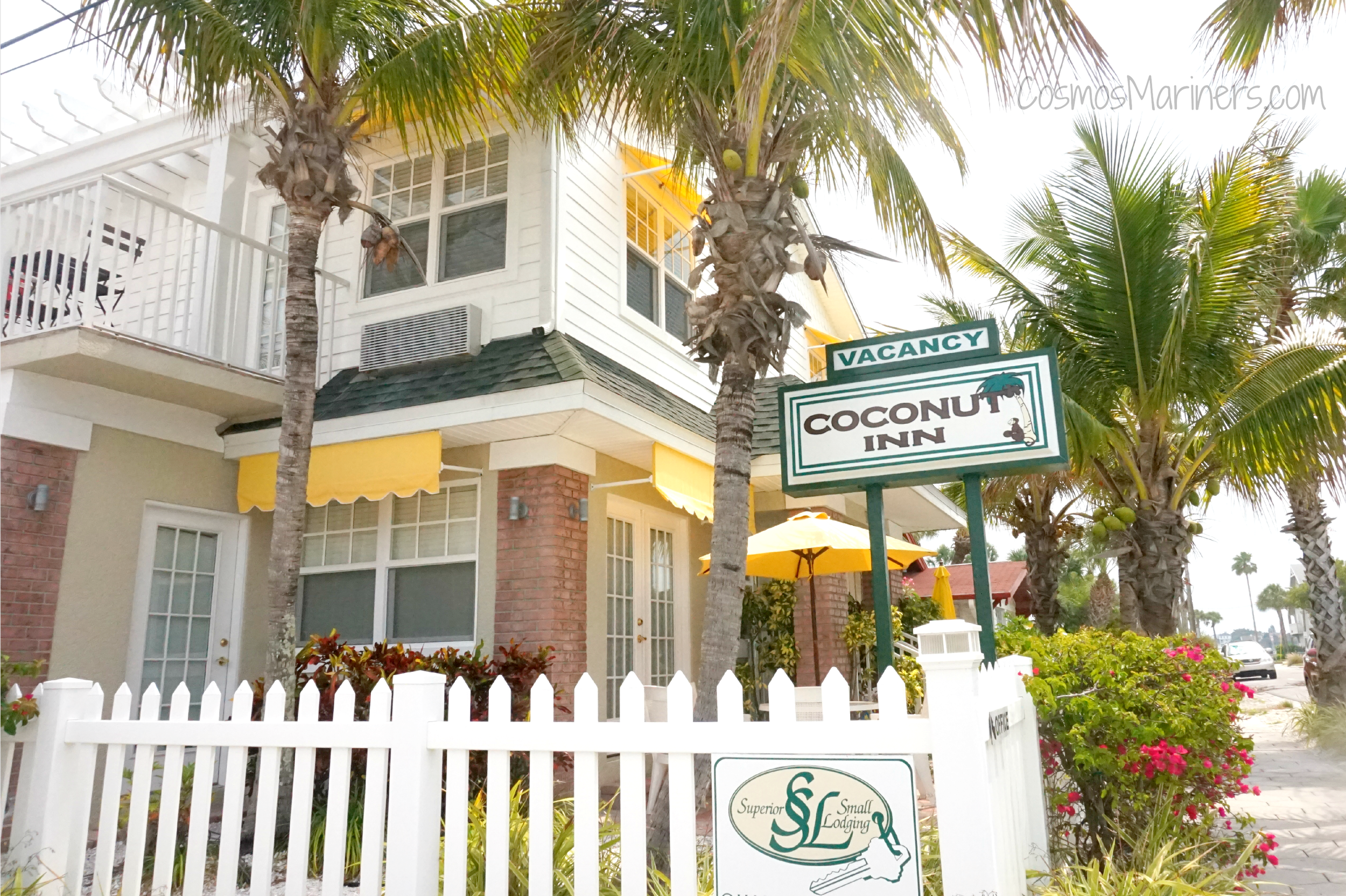 The white paneling and red brick exterior of the Coconut Inn in Pass-a-Grille, Florida, with a vacancy sign and palm trees in front