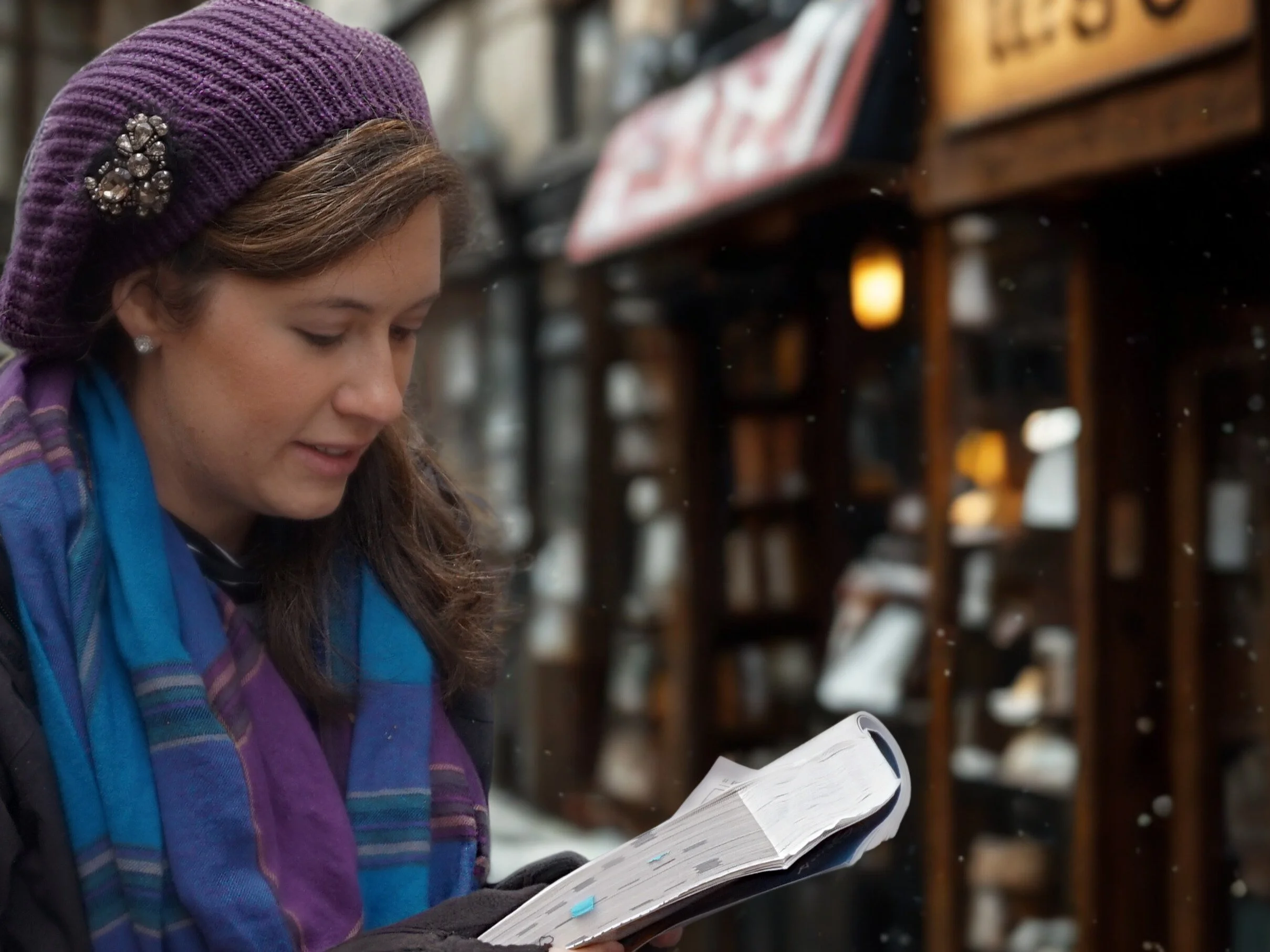 The author reading from a travel book in front of old stores in Munich and wearing a purple and blue plaid travel scarf