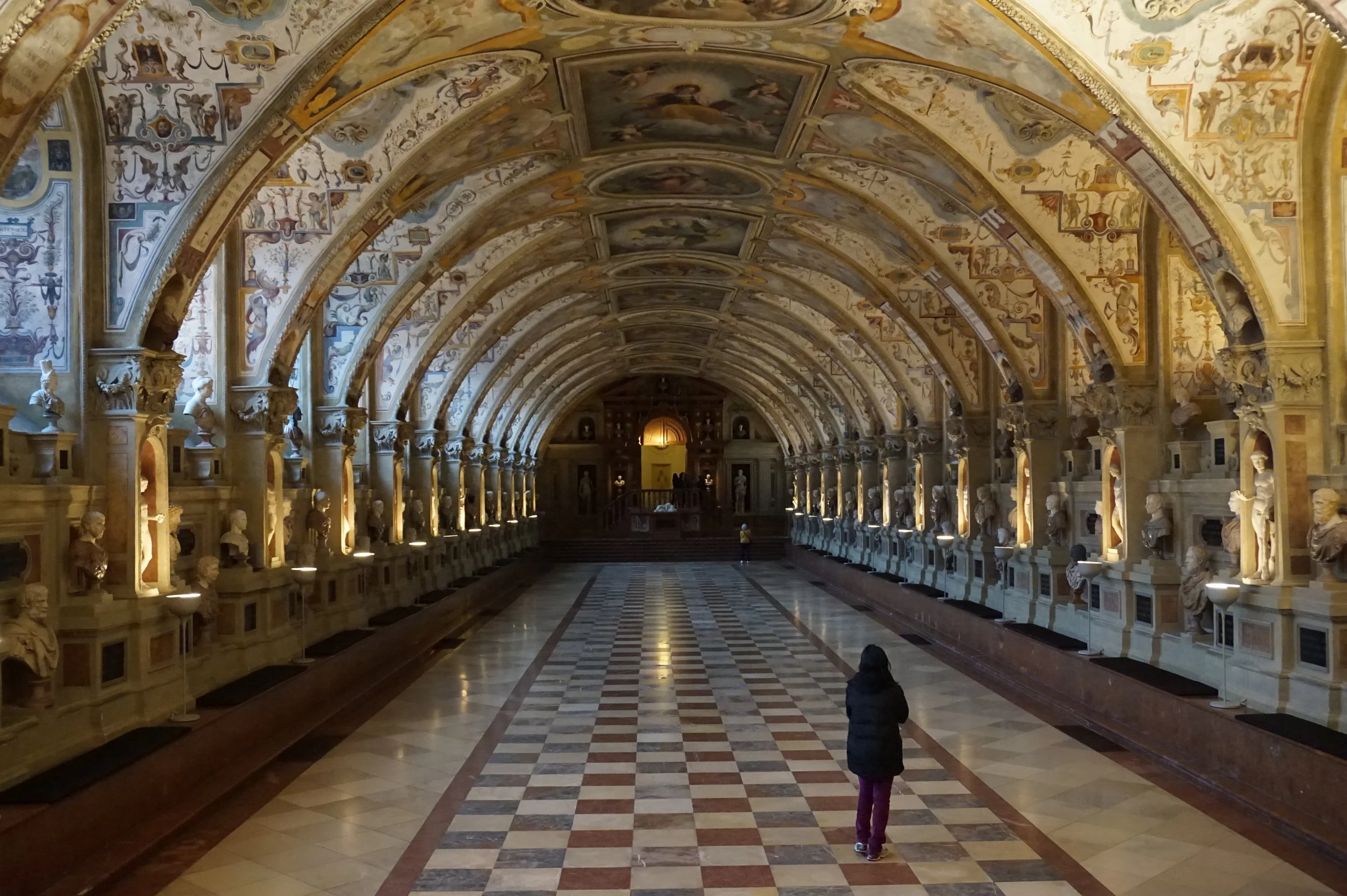 Woman in dark coat stands in a vaulted room with intricate paintings and sculptures