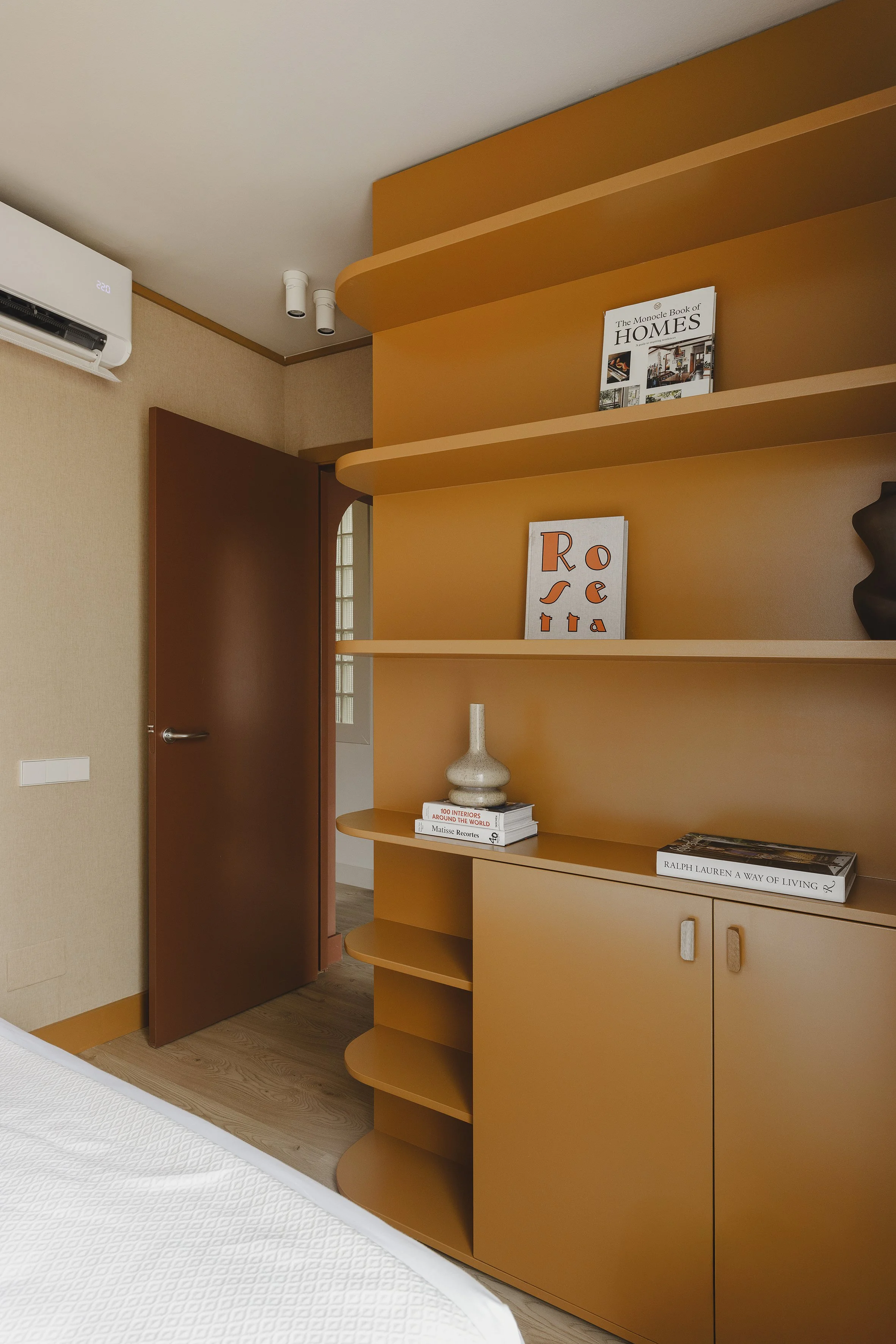 Interior de una habitación con una estantería de color mostaza con objetos decorativos y libros. En primer plano hay una cama parcialmente visible con ropa de cama blanca y una puerta de madera ligeramente abierta. En la pared hay un aparato de aire acondicionado.