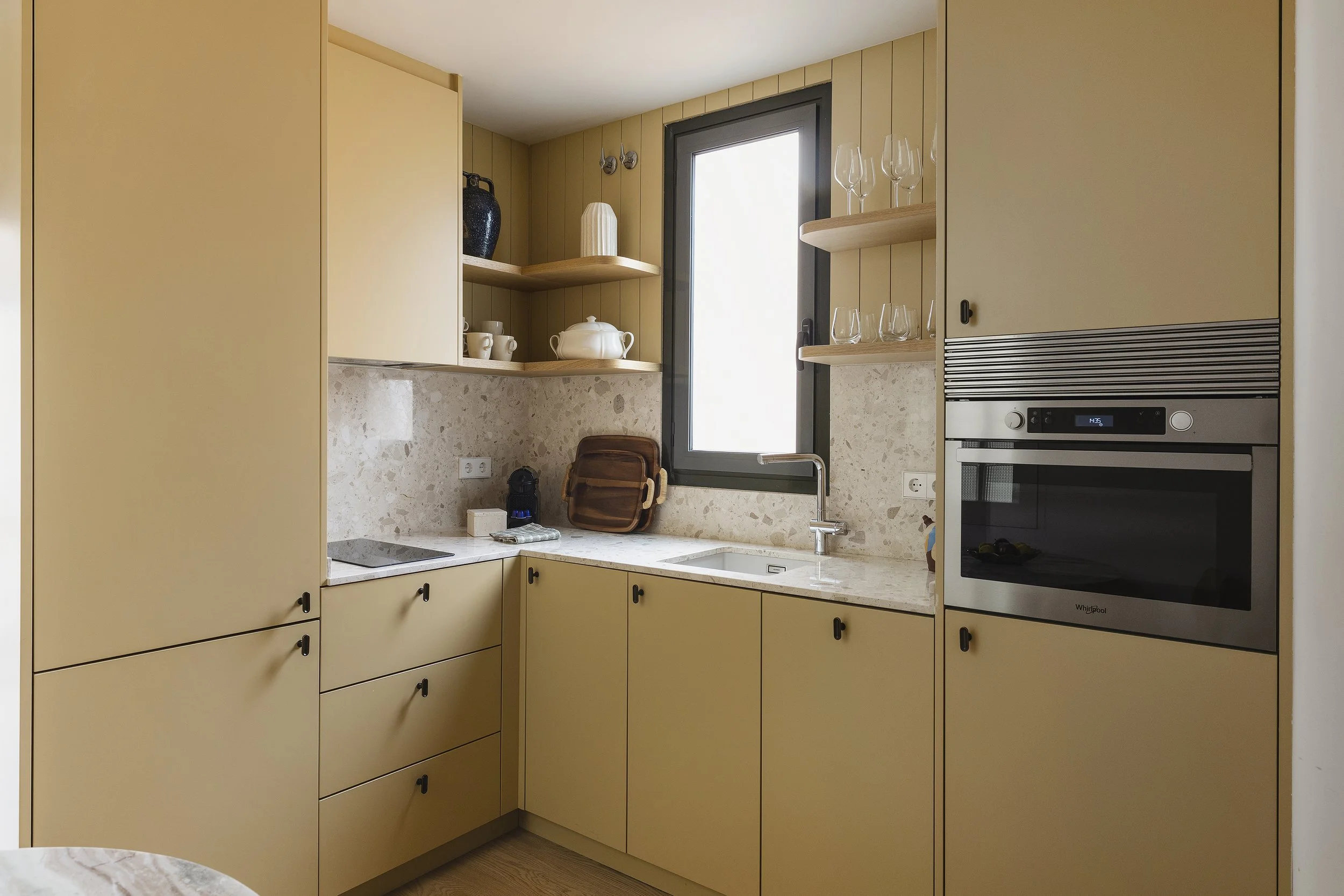 Una cocina moderna con armarios amarillos, estanterías abiertas y cerradas, una ventana y un horno empotrado. Las encimeras tienen una cafetera, una bolsa marrón y un dispensador de jabón. En los estantes hay cristalería, tazas y objetos decorativos.