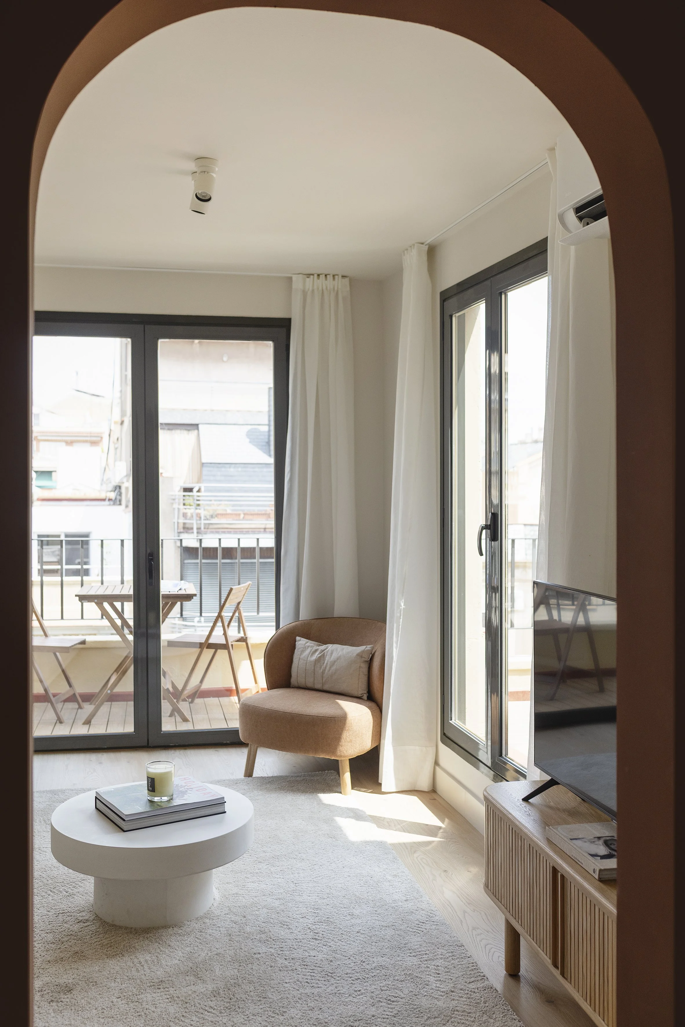 Salón con puerta corredera de cristal que da al balcón, sillón color canela con almohada, mesa de centro redonda blanca con libros y vela, alfombra beige, soporte de TV de madera con TV de pantalla plana, cortinas blancas y muebles de madera para el balcón.