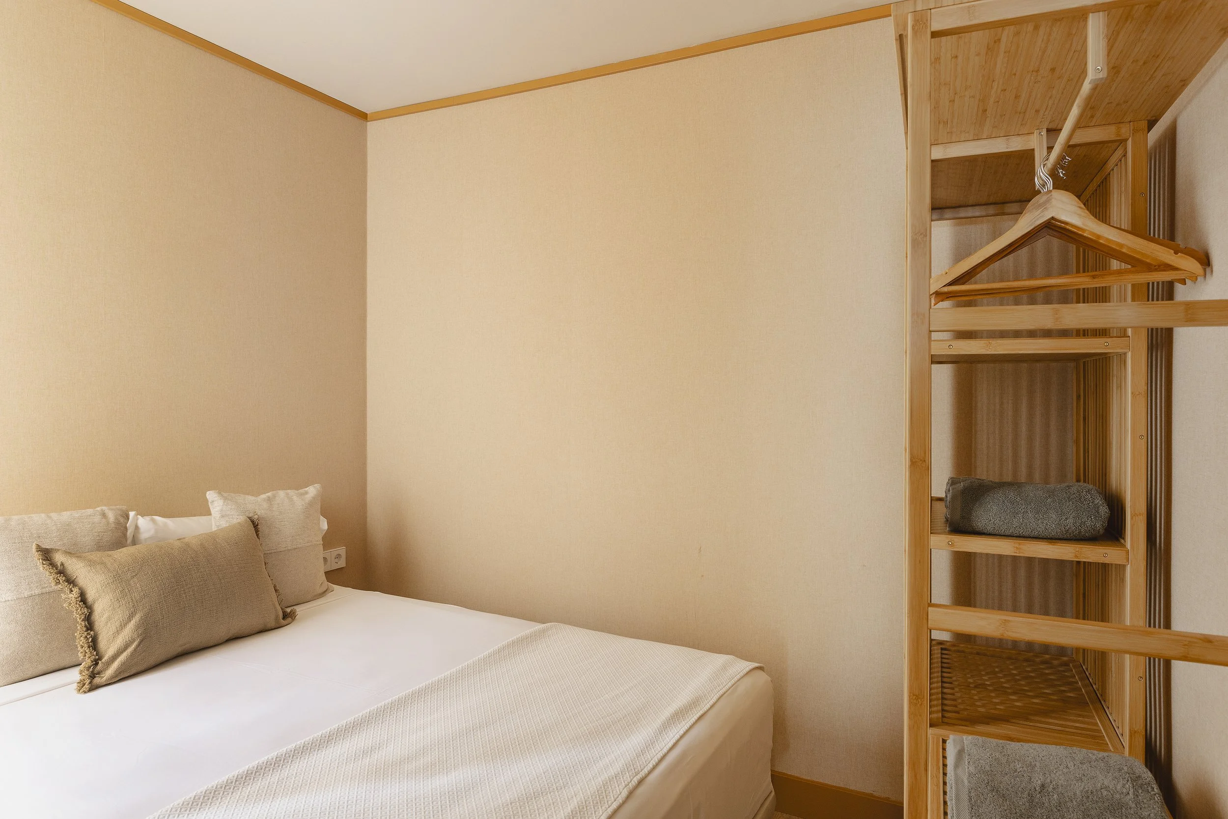 Una cama bien hecha con almohadas beige y una manta beige en un acogedor dormitorio con paredes de color claro y un armario esquinero con estantes de madera y espacio para colgar.
