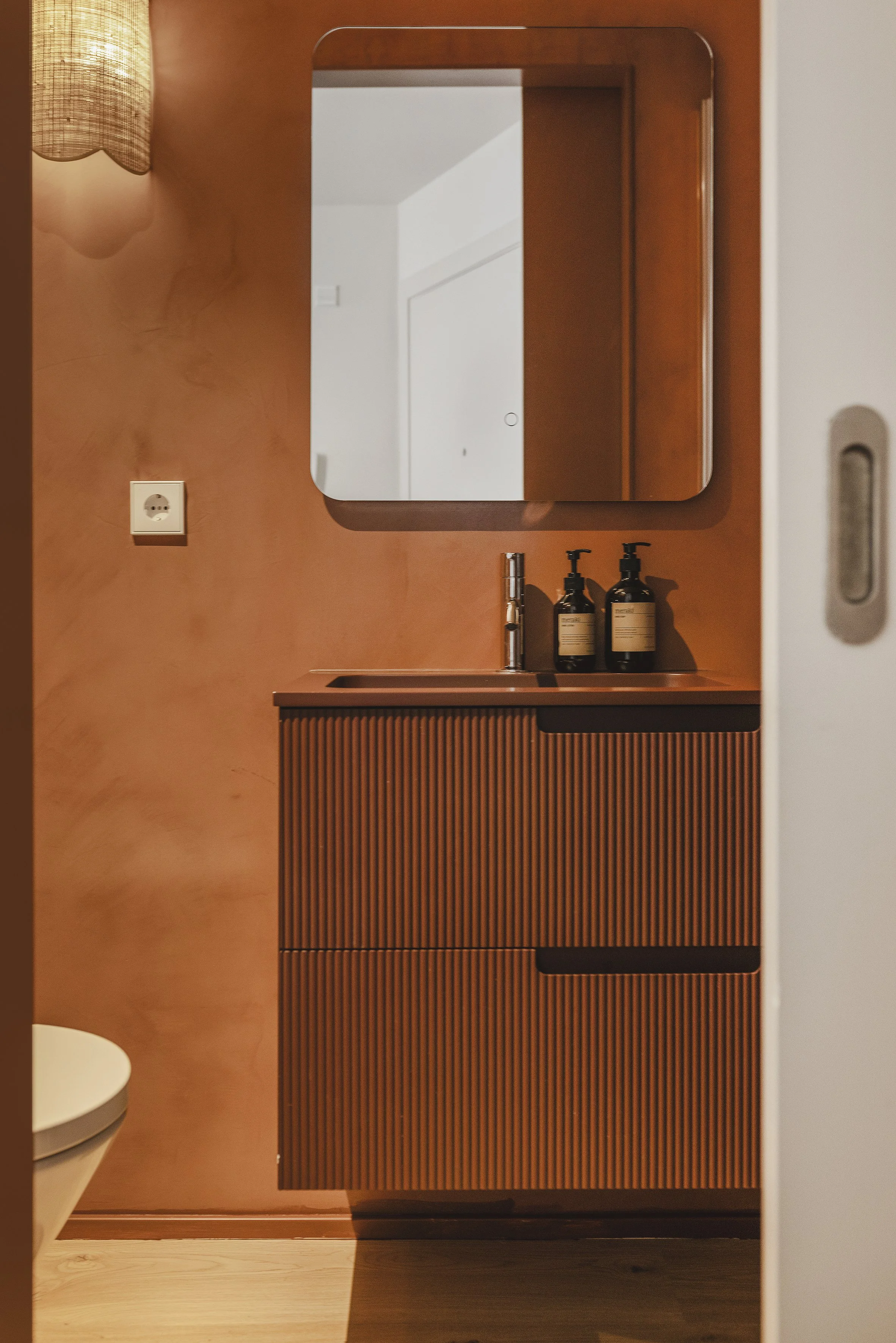 Un cuarto de baño con un tocador de madera, dosificadores de jabón negros, un espejo, una puerta de madera, un aplique y una pared blanca.