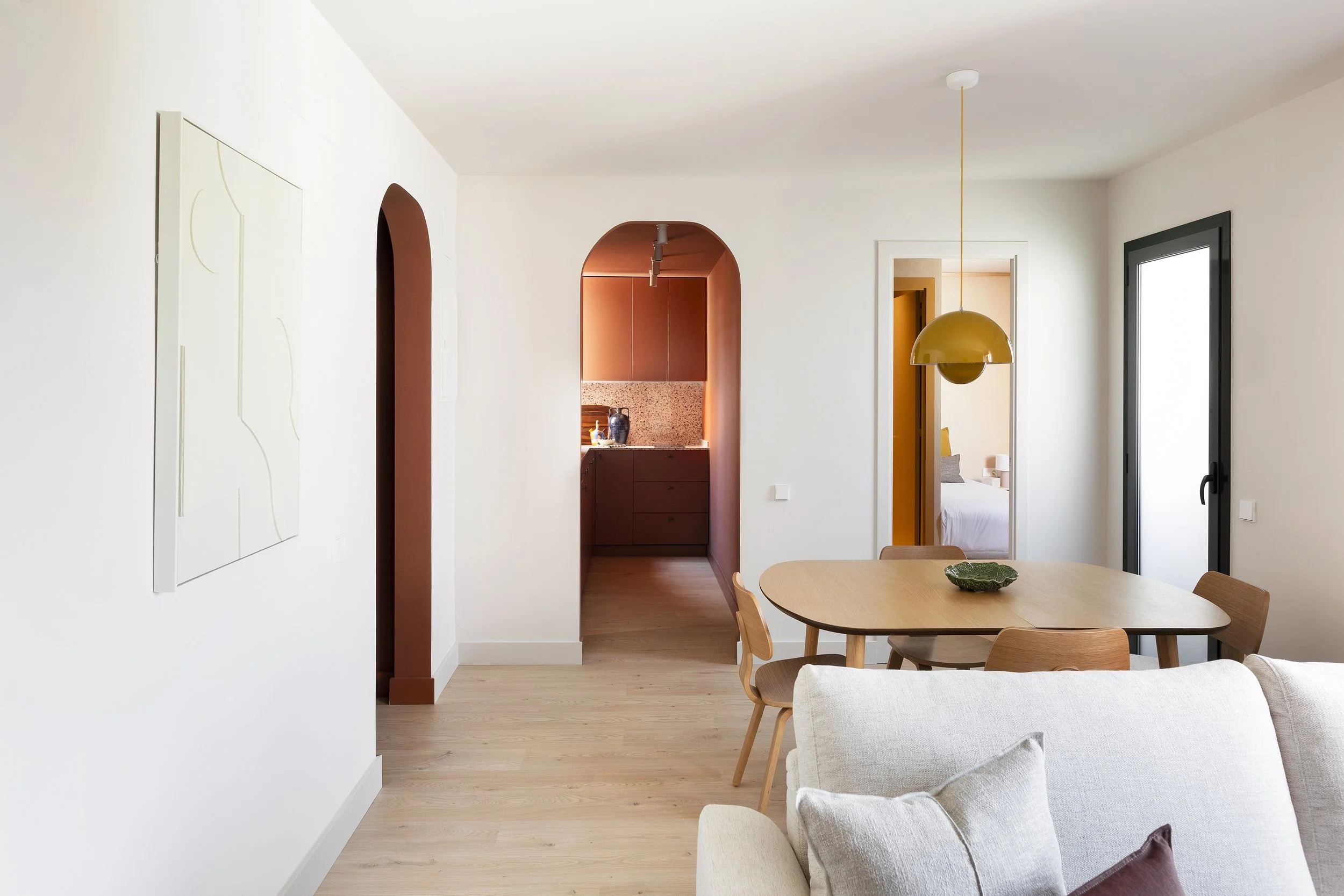 A bright living room with a dining area, white walls, light wood flooring, a white sofa with pillows, a wooden dining table with chairs, a yellow pendant light, and an arched doorway leading to a kitchen and bedrooms.