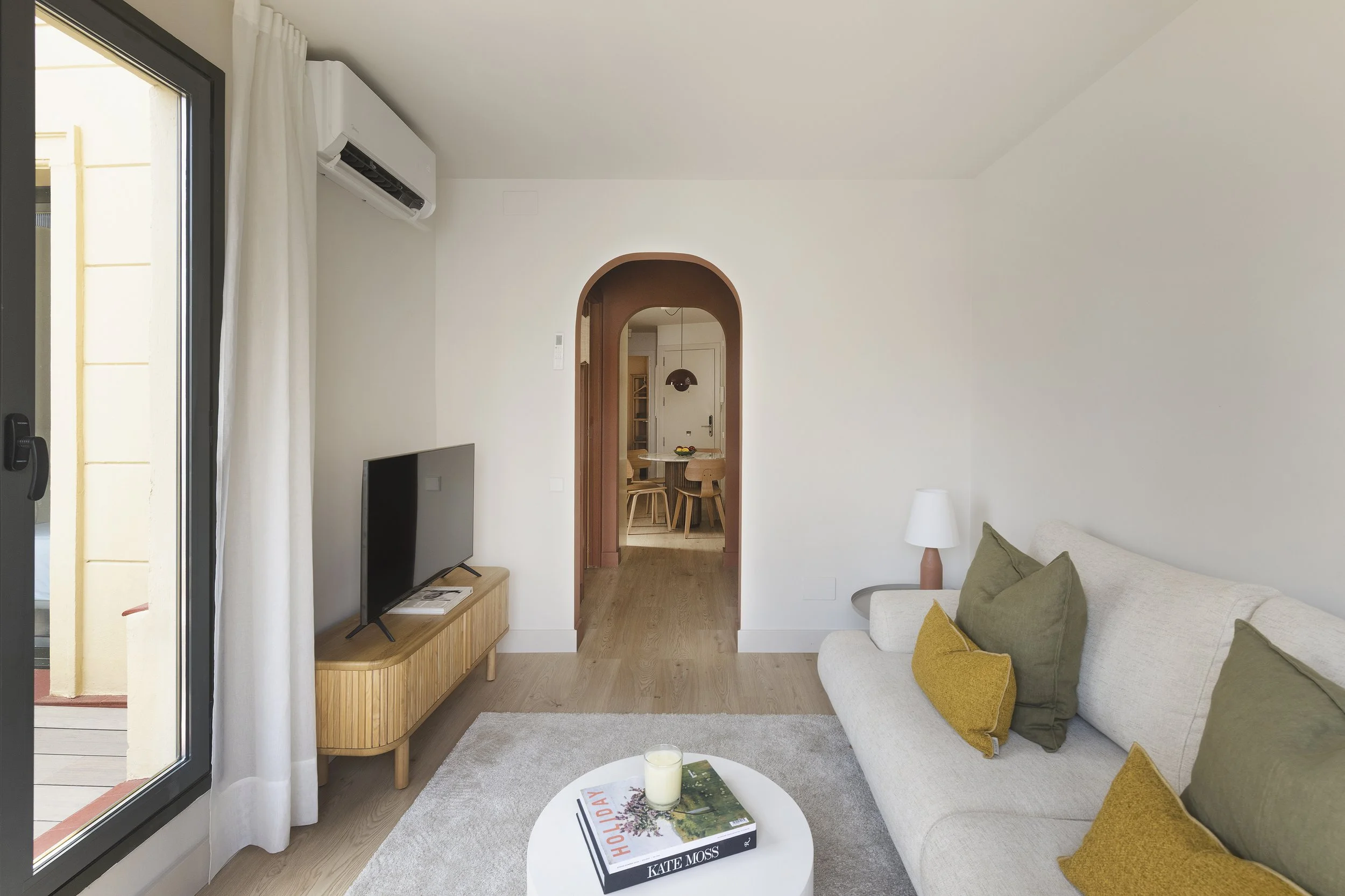 Sala de estar con un sofá blanco con cojines amarillos y verdes, una mesa de centro redonda blanca con un libro y una vela, un mueble de televisión de madera con un televisor de pantalla plana, una ventana con cortinas blancas y un aparato de aire acondicionado, con una puerta arqueada que da a un