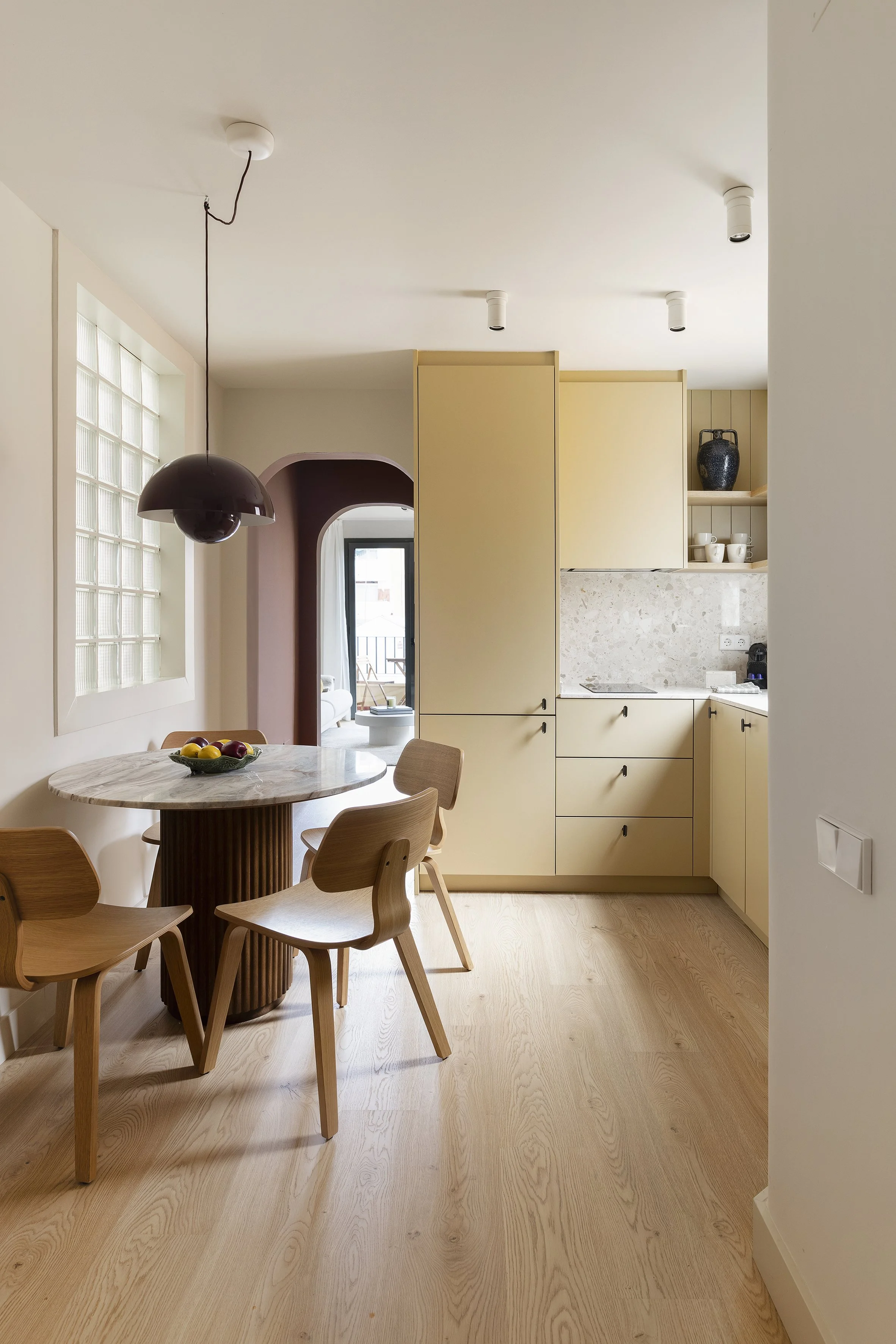 Vista interior de una cocina y comedor con una mesa redonda de madera, cuatro sillas de madera, armarios de cocina amarillos, salpicadero de mármol y una gran ventana con bloques de cristal.