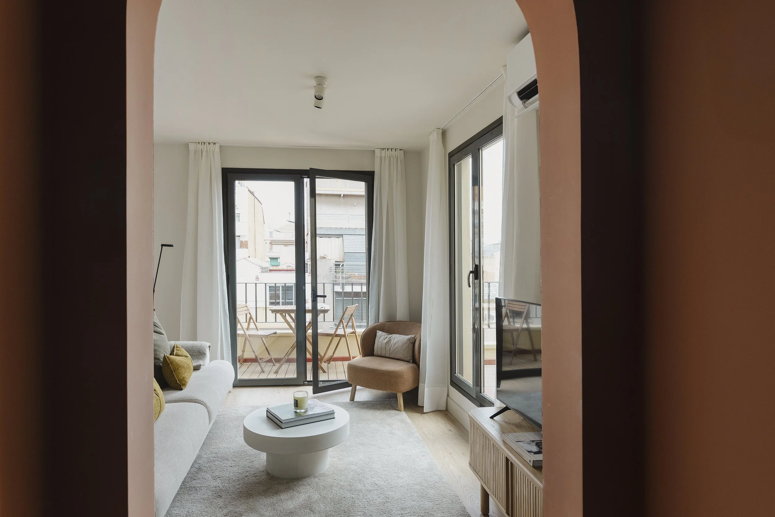 Salón con sofá blanco, sillón beige, mesa de centro blanca, puertas correderas de cristal al balcón y televisión.