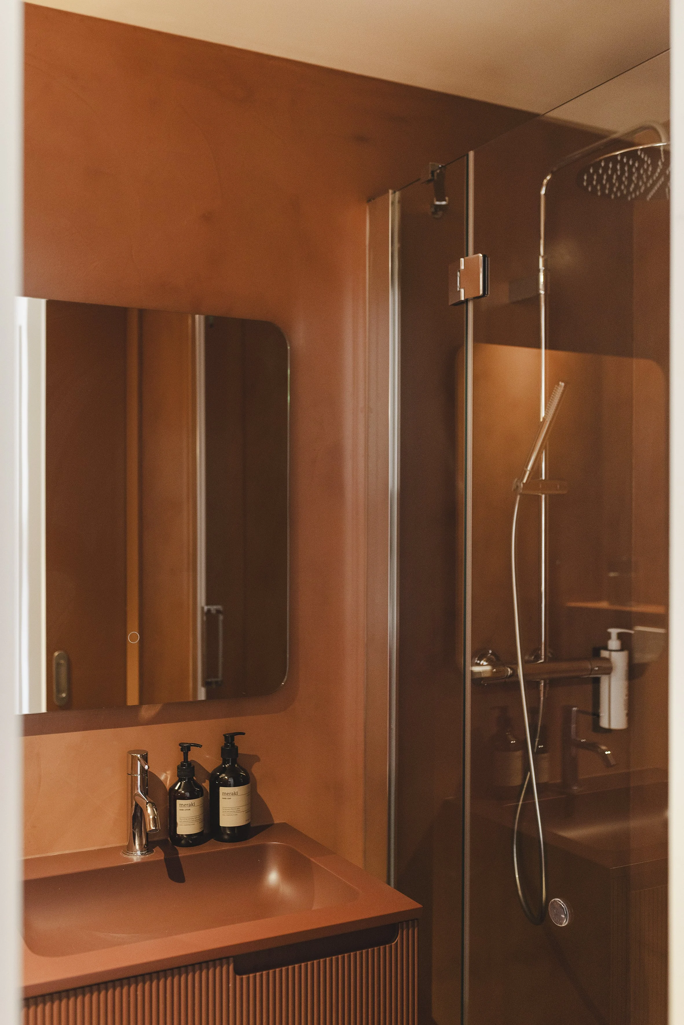 Un cuarto de baño con un espejo montado en la pared sobre un lavabo, dispensadores de jabón y una ducha con puertas de cristal transparente y cabezal de ducha manual.