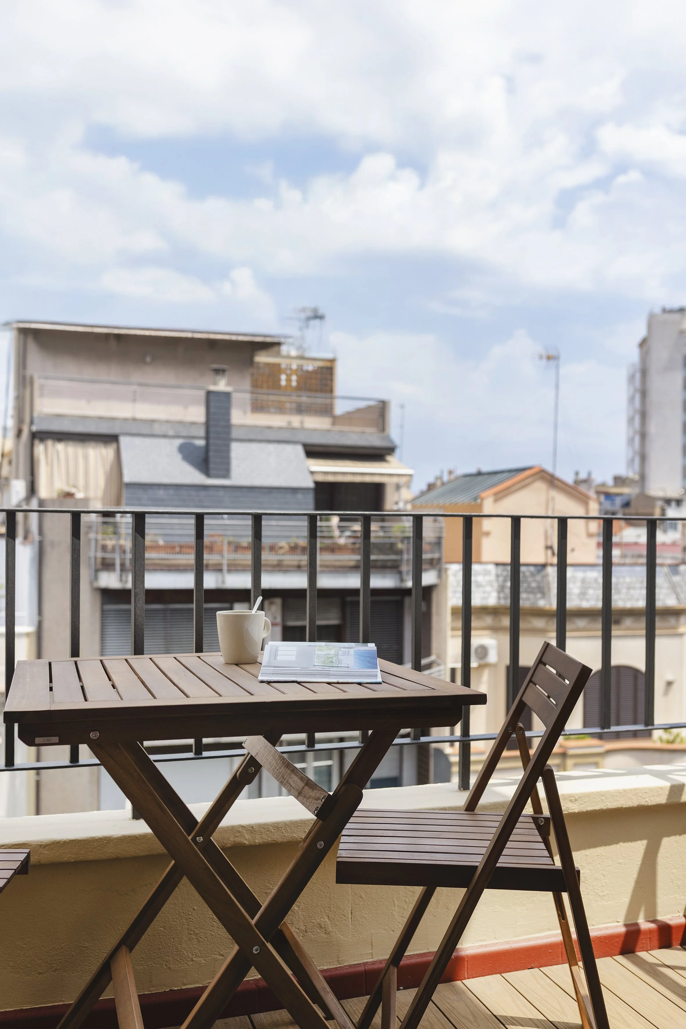 Balcón con mesa y silla de madera, taza con cuchara, revista abierta, con vistas a los edificios vecinos bajo un cielo parcialmente nublado.