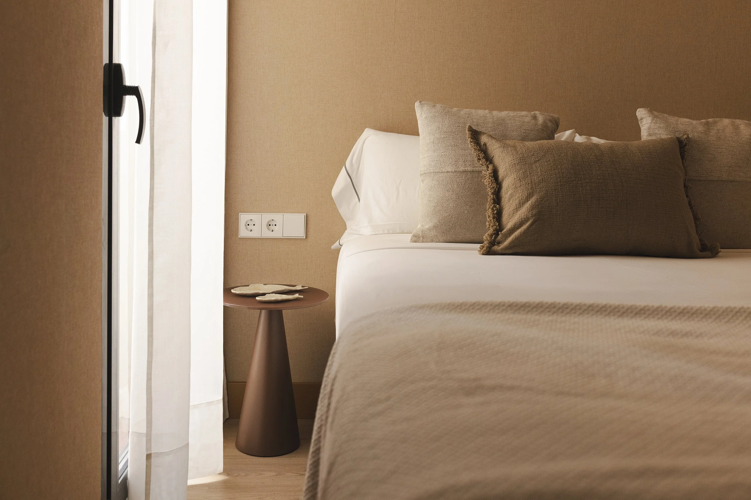 Primer plano de una cama bien hecha con almohadas beige y cabecero marrón en una habitación de hotel, con una mesita auxiliar marrón y una ventana con cortinas blancas al lado.