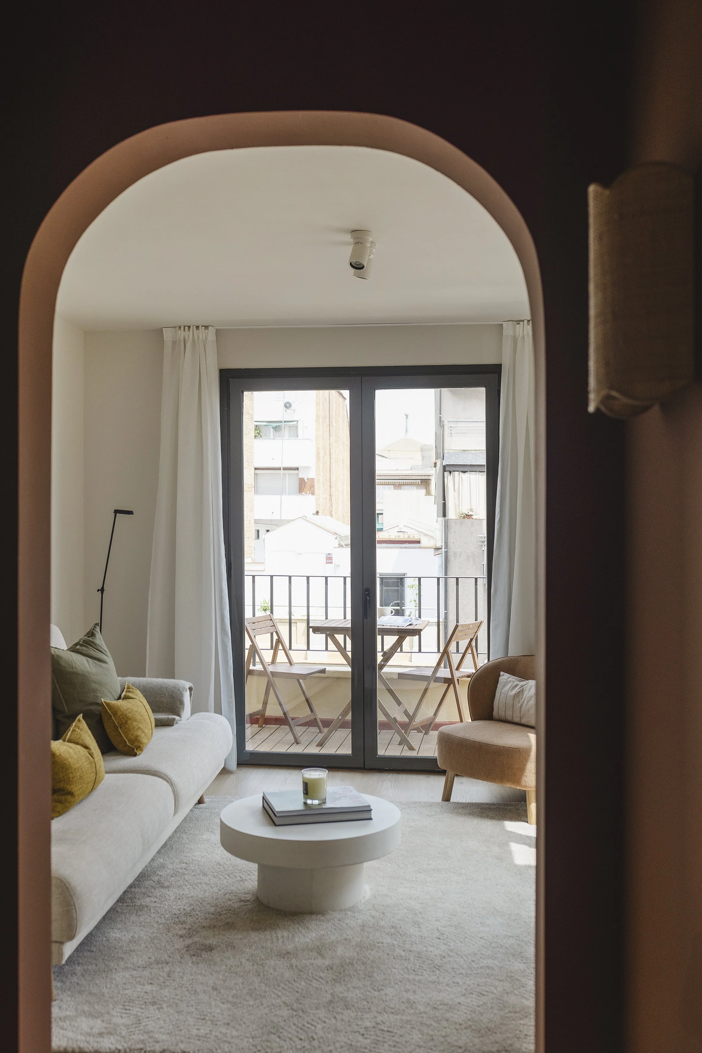 Sala de estar con sofá, sillón, mesa de centro con libros y una vela, y un balcón con una mesa y dos sillas al aire libre.