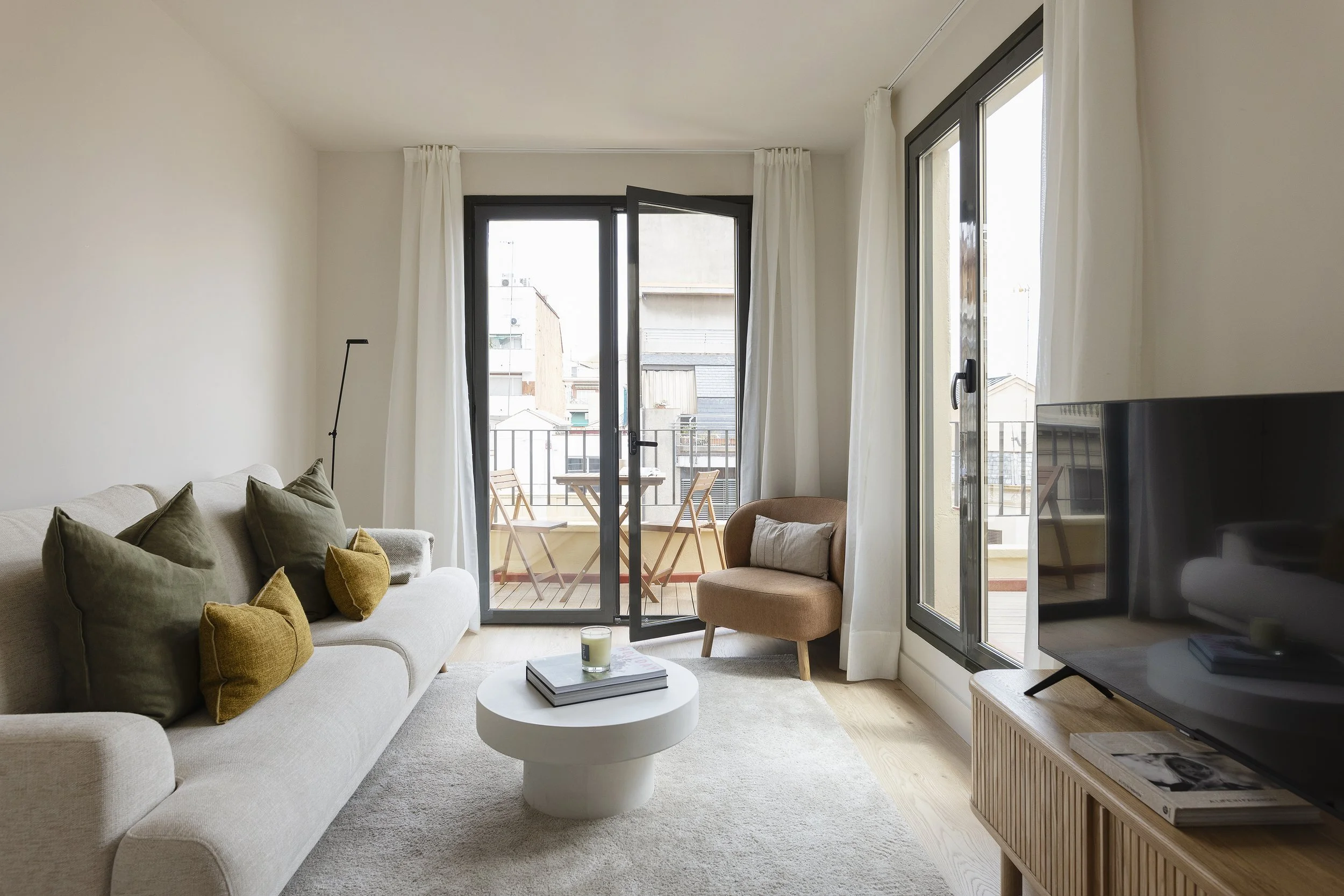 Sala de estar con un sofá beige, cojines marrones y mostaza, una mesa de centro blanca redonda, un sillón rosa con almohada, un televisor de pantalla plana grande sobre un soporte de madera y puertas correderas de cristal que dan a un balcón con una mesa pequeña y dos sillas.