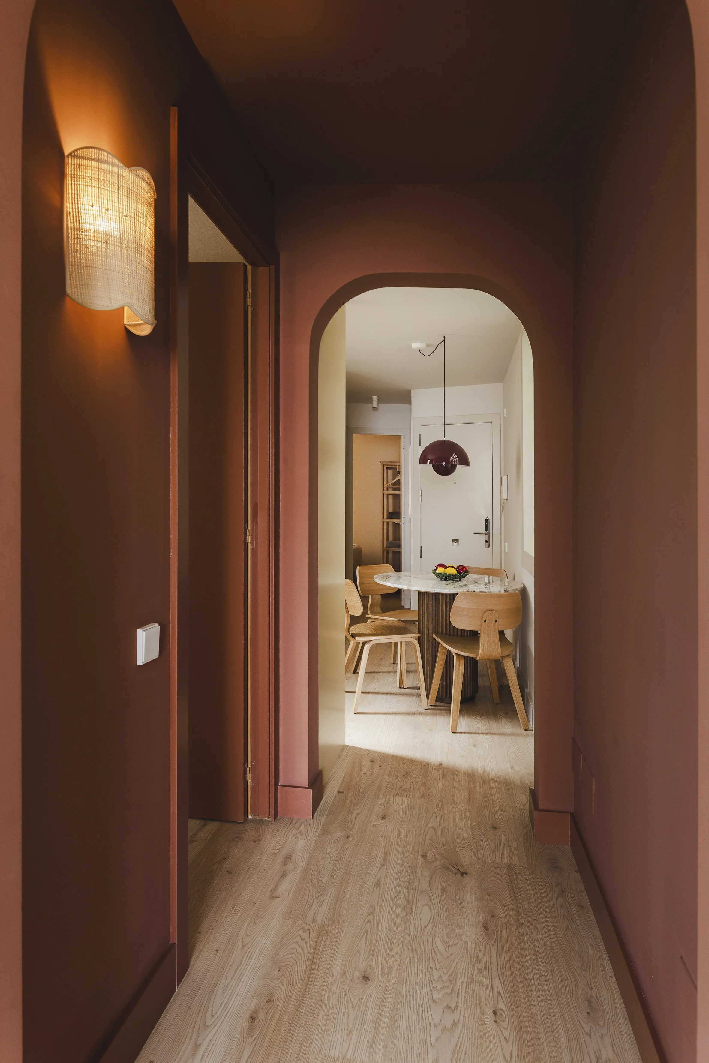 Un pasillo con paredes de color rosa oscuro y suelo de madera clara que conduce a una zona de comedor con una mesa redonda de mármol y cuatro sillas de madera. Hay una lámpara colgante rojo oscuro colgando sobre la mesa y un frutero encima. En el fondo se ve la puerta de entrada.
