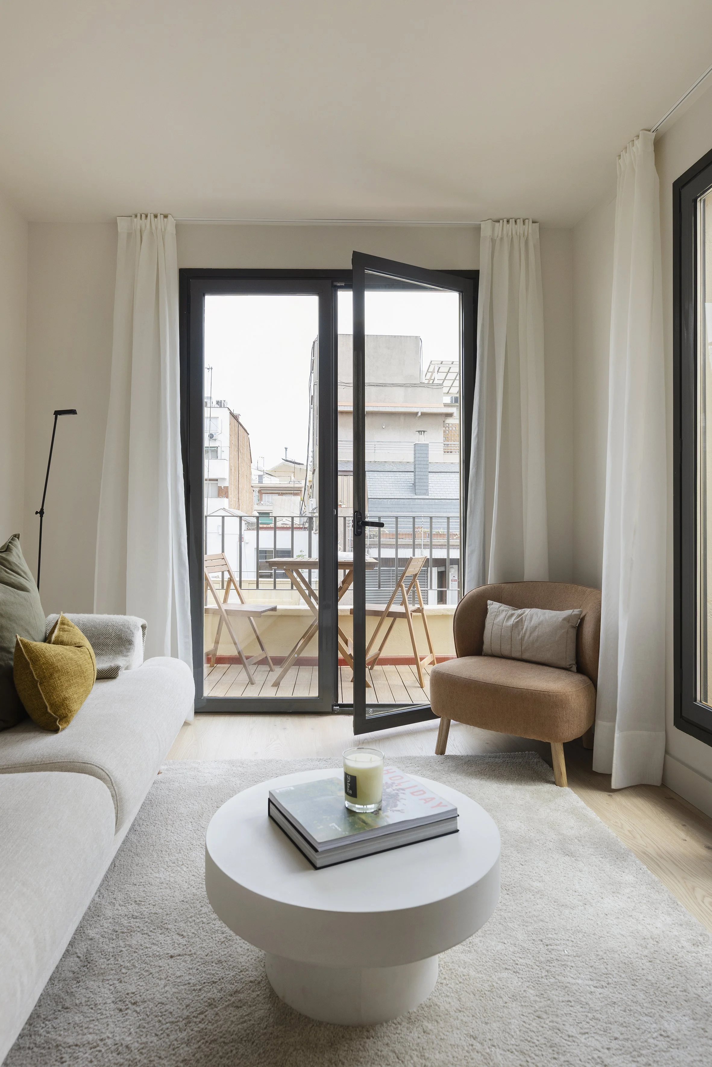 Salón con un sofá blanco, un sillón beige, una mesa de centro con una vela y libros, y un balcón con dos sillas y una mesa visible a través de puertas de cristal.