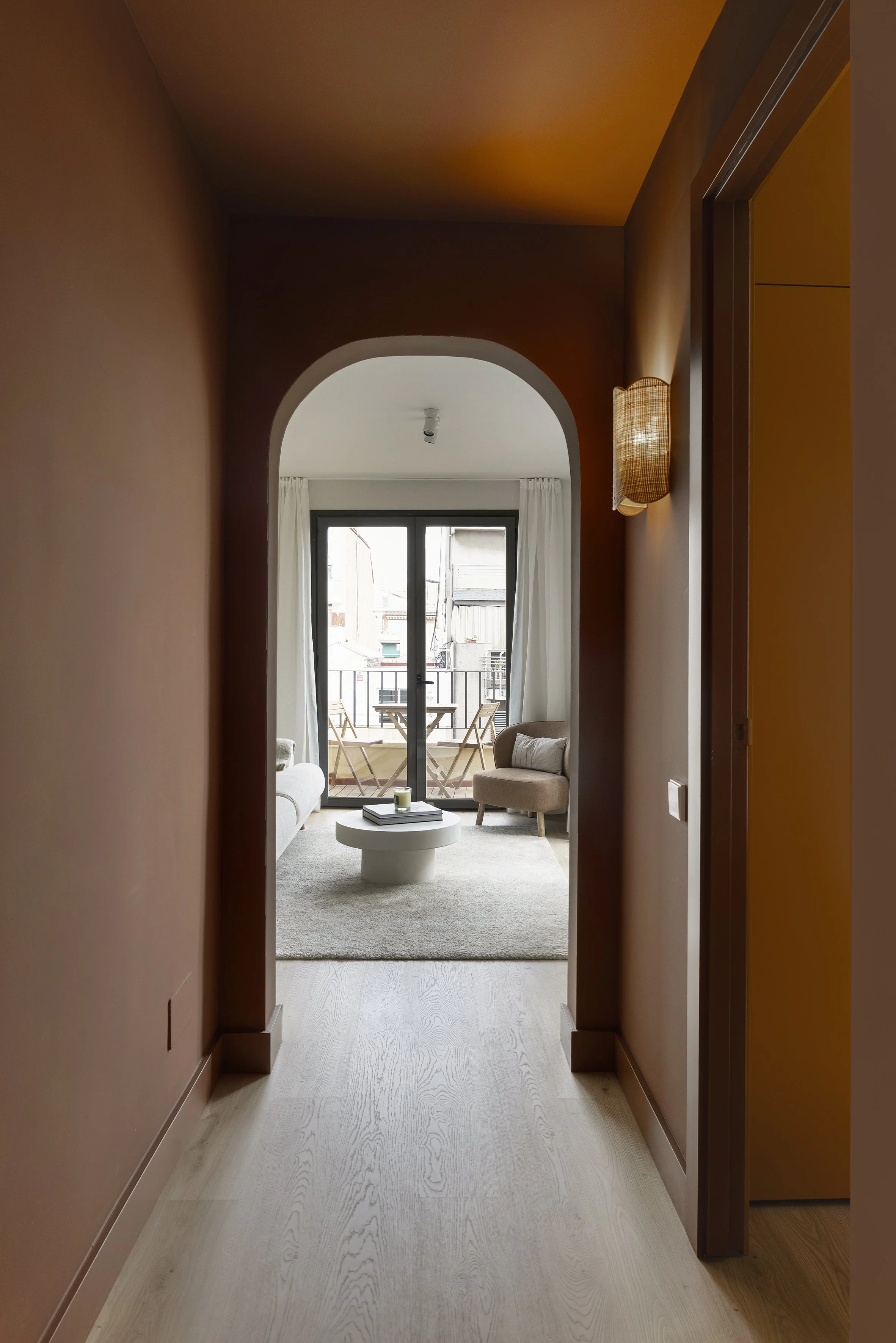 Vista a través de una puerta arqueada a un salón con puertas correderas de cristal que dan a un balcón, amueblado con un sillón beige, un sofá blanco, una mesa de centro redonda y una pequeña mesa de exterior con sillas.
