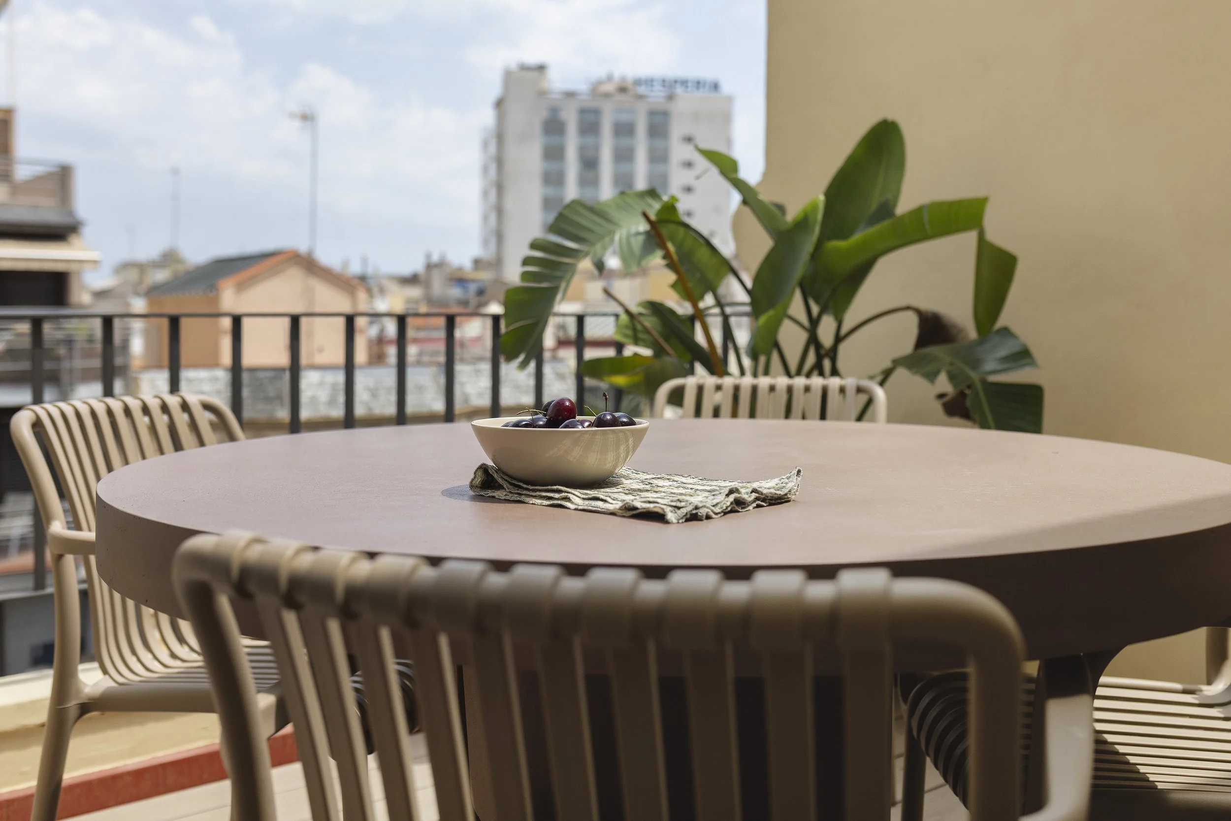 Balcón al aire libre con una mesa redonda y cuatro sillas de color beige, un tazón de cerezas en una servilleta, gran planta de interior verde, edificios de la ciudad en el fondo.