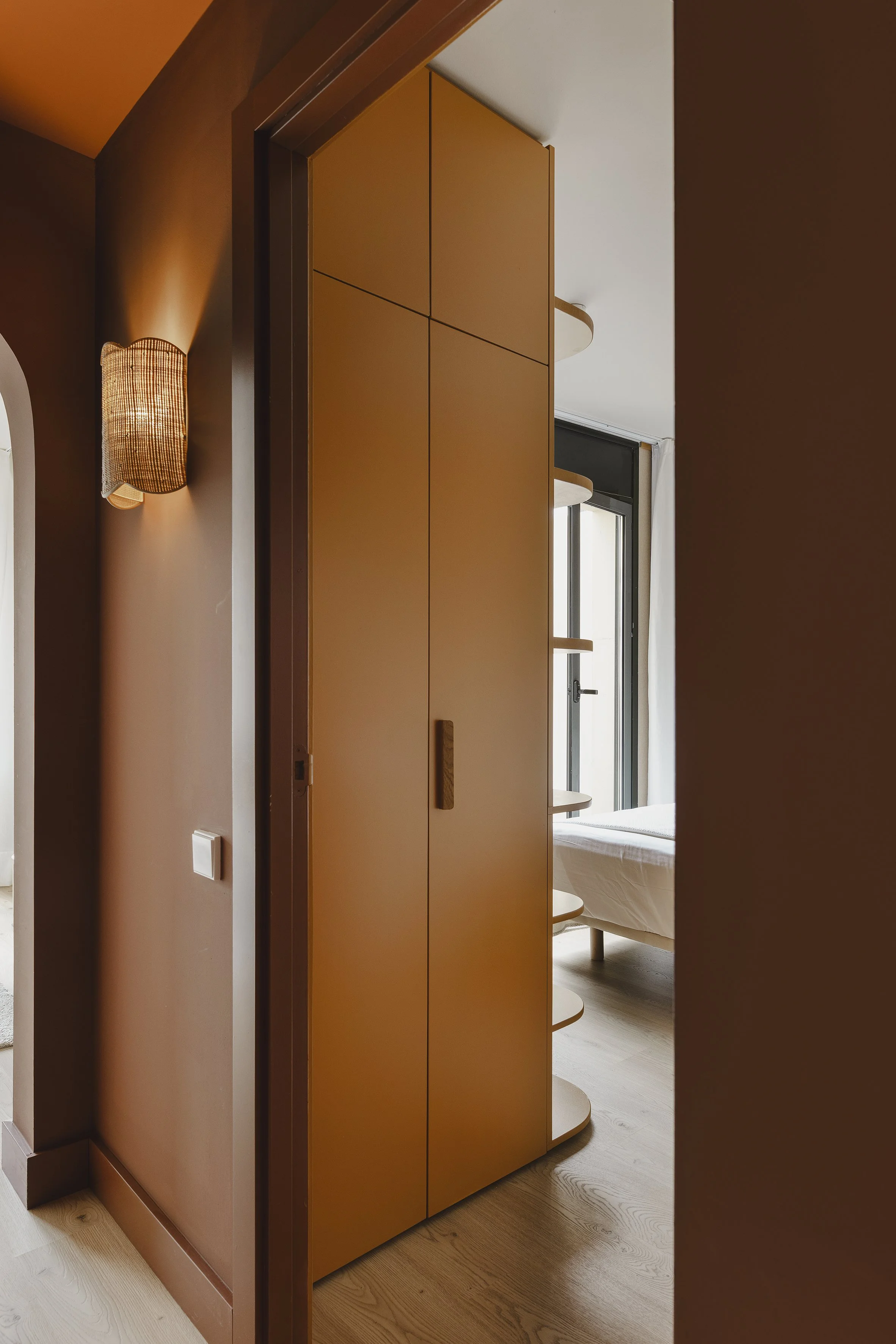 Vista de una habitación interior parcialmente oscurecida por una puerta corredera con estanterías, una cama cerca de una ventana y una lámpara de pared.