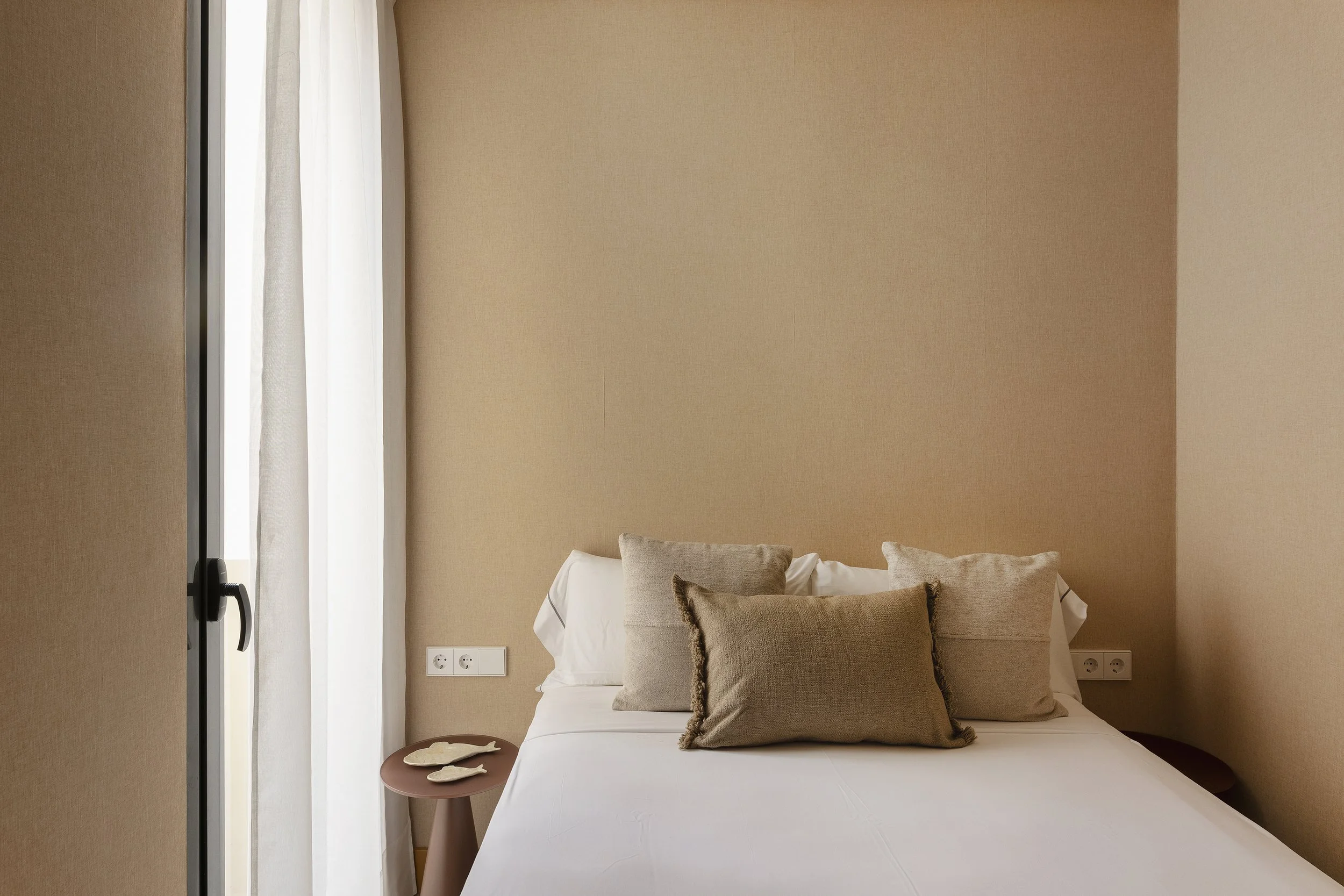 Una cama bien hecha con almohadas beige y blancas en una habitación con paredes beige y una ventana con cortinas blancas.