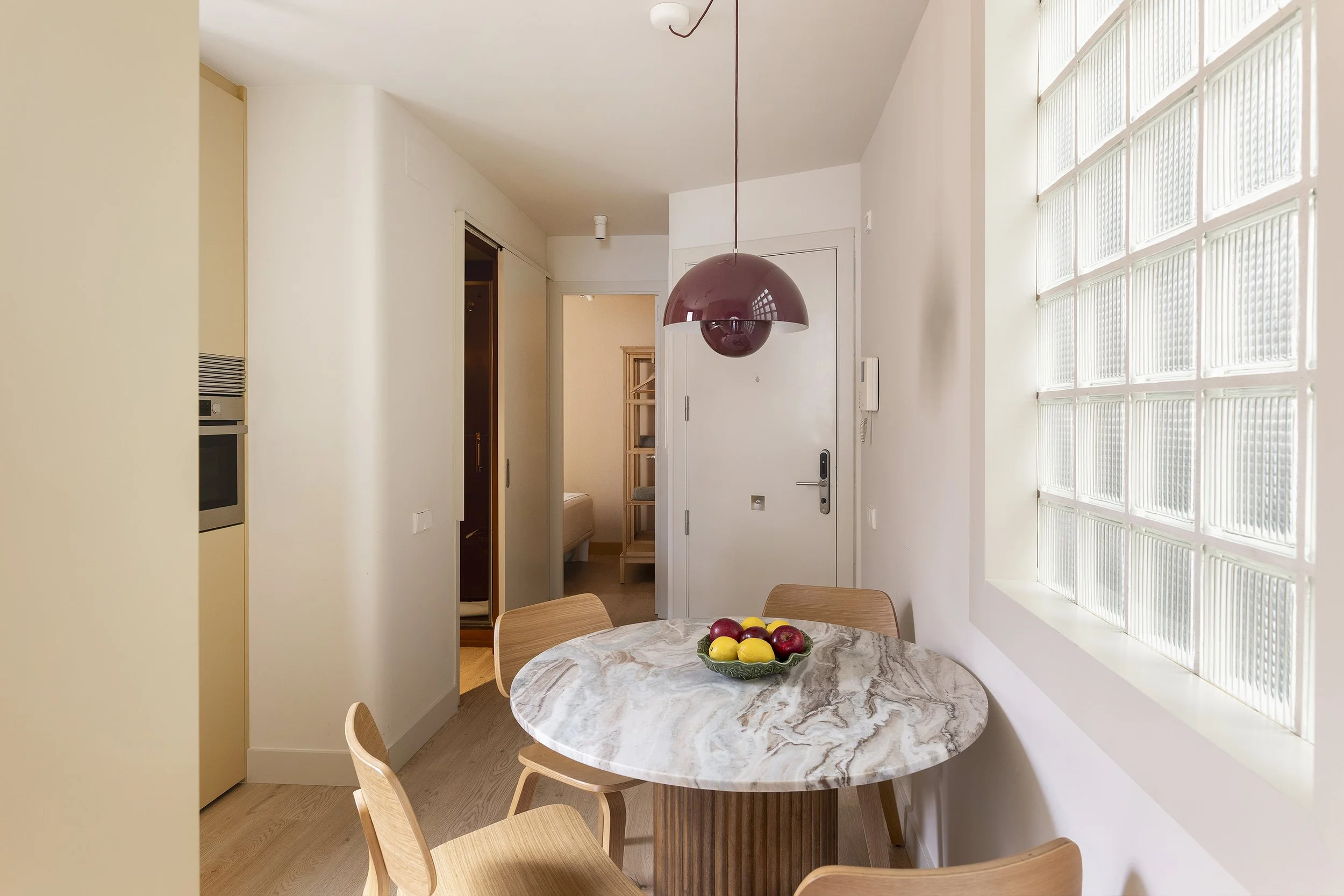 Un pequeño comedor con una mesa redonda de mármol y cuatro sillas de madera. Sobre la mesa hay un cuenco con manzanas y limones. Hay una gran ventana con bloques de cristal a la derecha y una puerta blanca al fondo. Sobre la mesa cuelga una lámpara colgante de color granate. El sitio